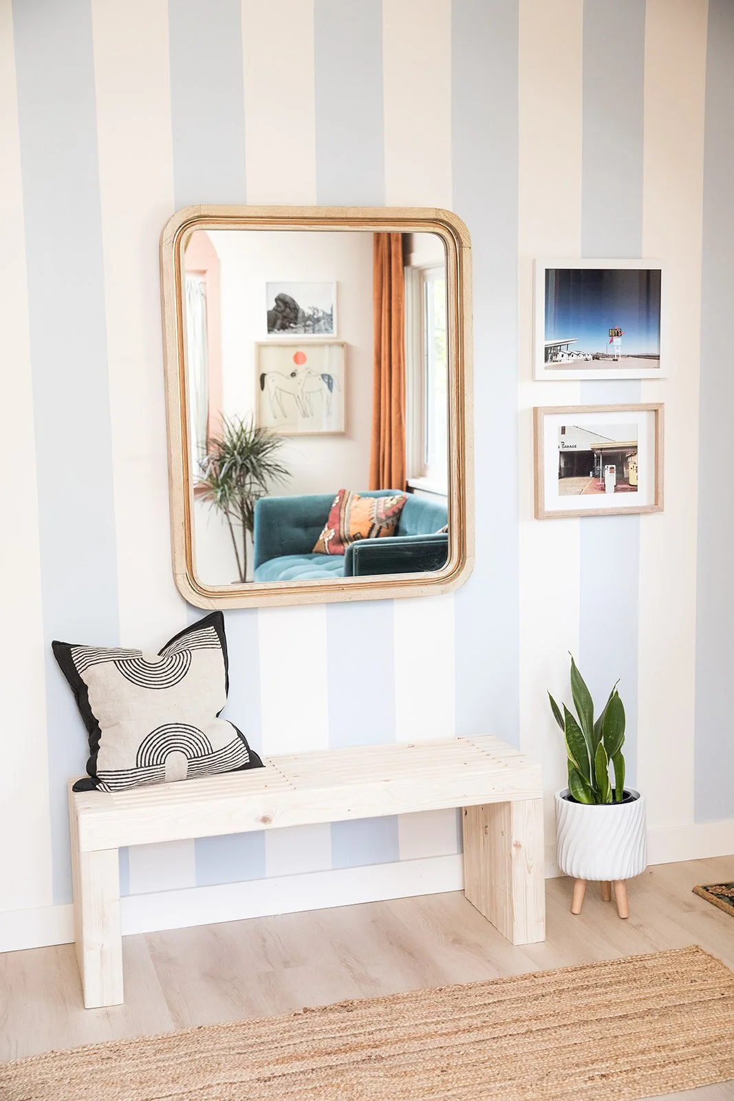 A room with a white bench, a patterned pillow, a potted plant, a wooden-framed mirror, and three framed photos on a striped wall.