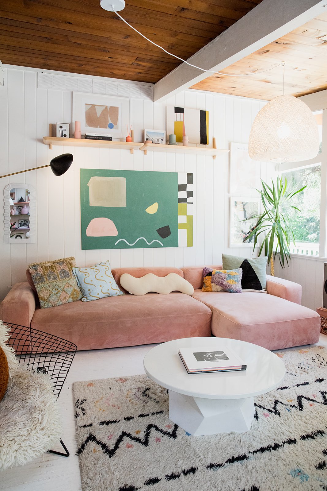 Living room with pink sofa, abstract art on white paneled wall, green houseplant near window, white coffee table with magazines, patterned pillows, woven pendant light, and wooden ceiling.