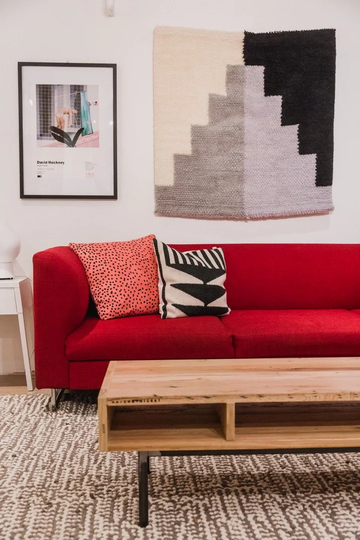 Living room with a red sofa, two decorative pillows (one pink with black dots and one white with black abstract leaf design), a light wood coffee table, a textured black, white, and beige wall hanging, a framed art print of a person with a dog by a window, a white side table with a white lamp, and a patterned beige carpet.
