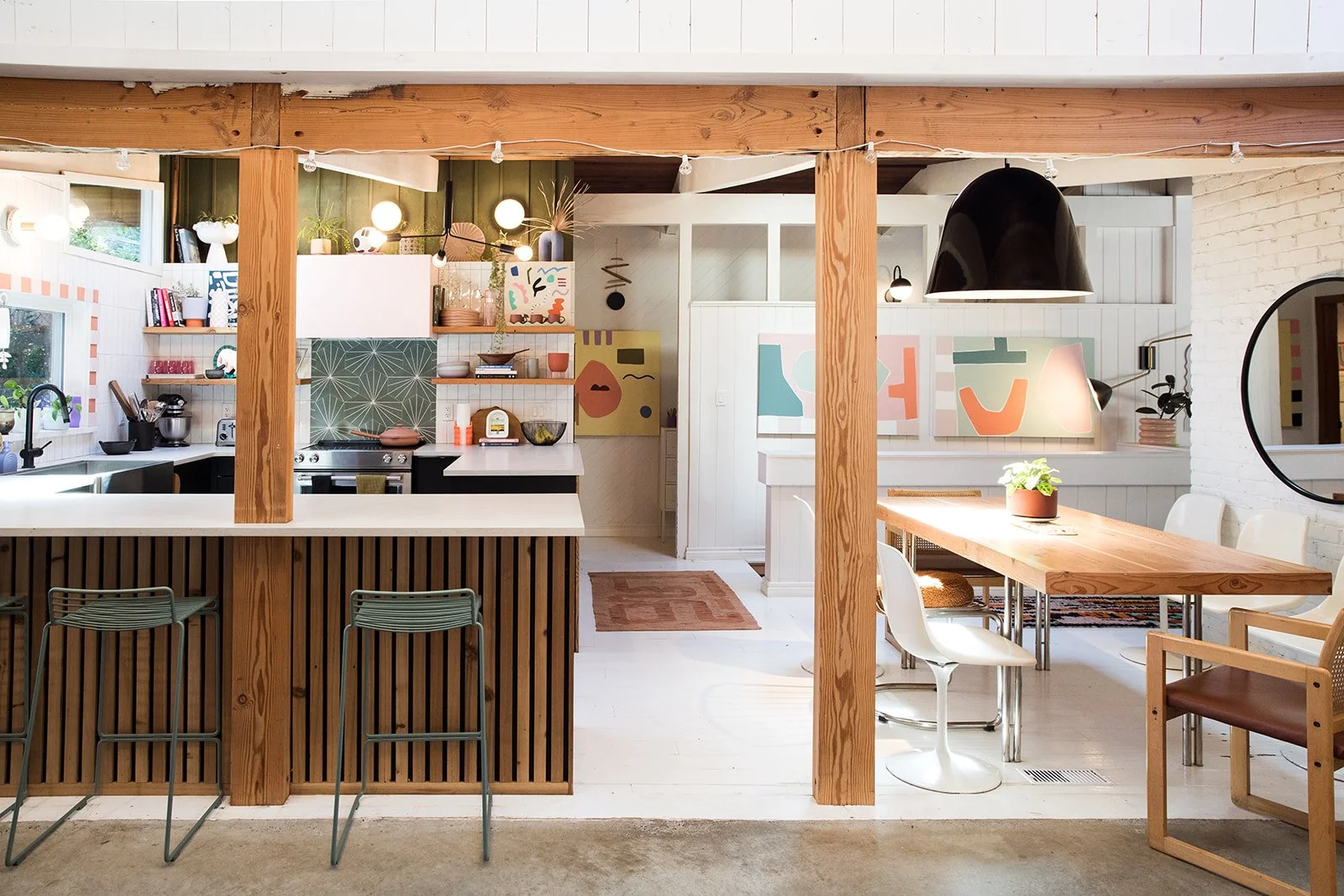 Bright, modern kitchen and dining area with colorful abstract art on white walls, wooden beams, and a long wooden dining table with white and tan chairs, illuminated by a large black pendant light.
