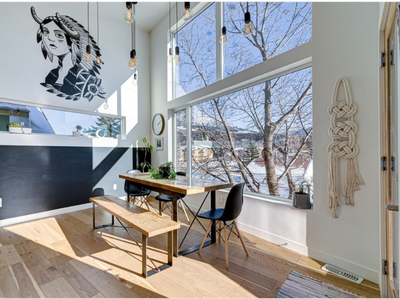 Bright dining area with large windows overlooking a snowy, tree-filled landscape, minimal decor, wall art, a wooden table with black chairs, a bench, and natural sunlight.