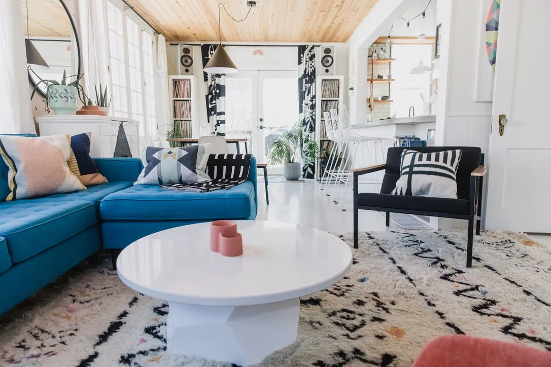 Bright living room with blue L-shaped sofa, black armchair, white coffee table, patterned rug, and large windows with curtains. Open kitchen area in background with bar stools, plants, and wooden ceiling.