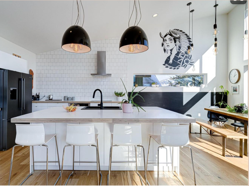 Modern kitchen with white cabinets, a large island with four white chairs, black pendant lights, a black refrigerator, and a wall with a black and white illustration of a woman with horns and feathers.