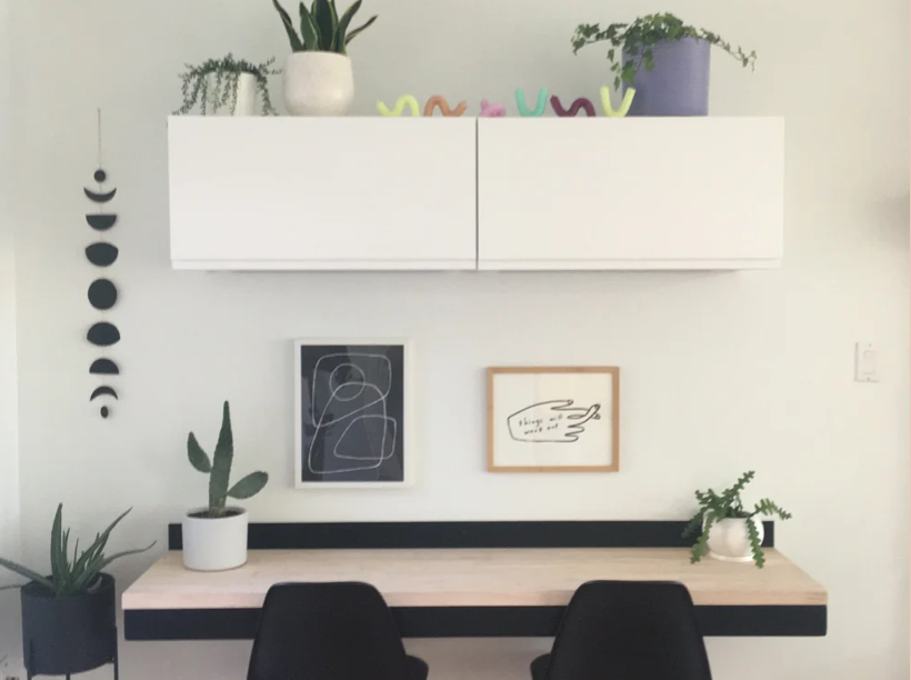 Minimalist home office with white wall, black desk, and two black chairs. Decor includes framed artwork, potted succulents, and hanging moon phase mobile.