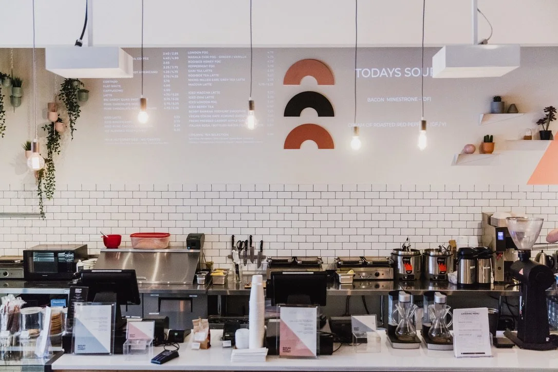 Interior of a modern cafe with a white tiled wall, food and drink menu, pendant lights, and a counter with coffee-making equipment.