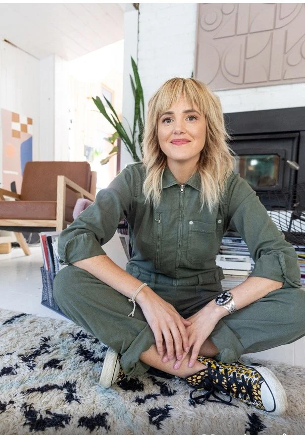 Young woman Interior designer with blonde hair sitting cross-legged on a rug indoors, smiling, wearing a green jumpsuit, patterned sneakers, a watch, and bracelet, with books and a plant in the background. Cool modern designer.