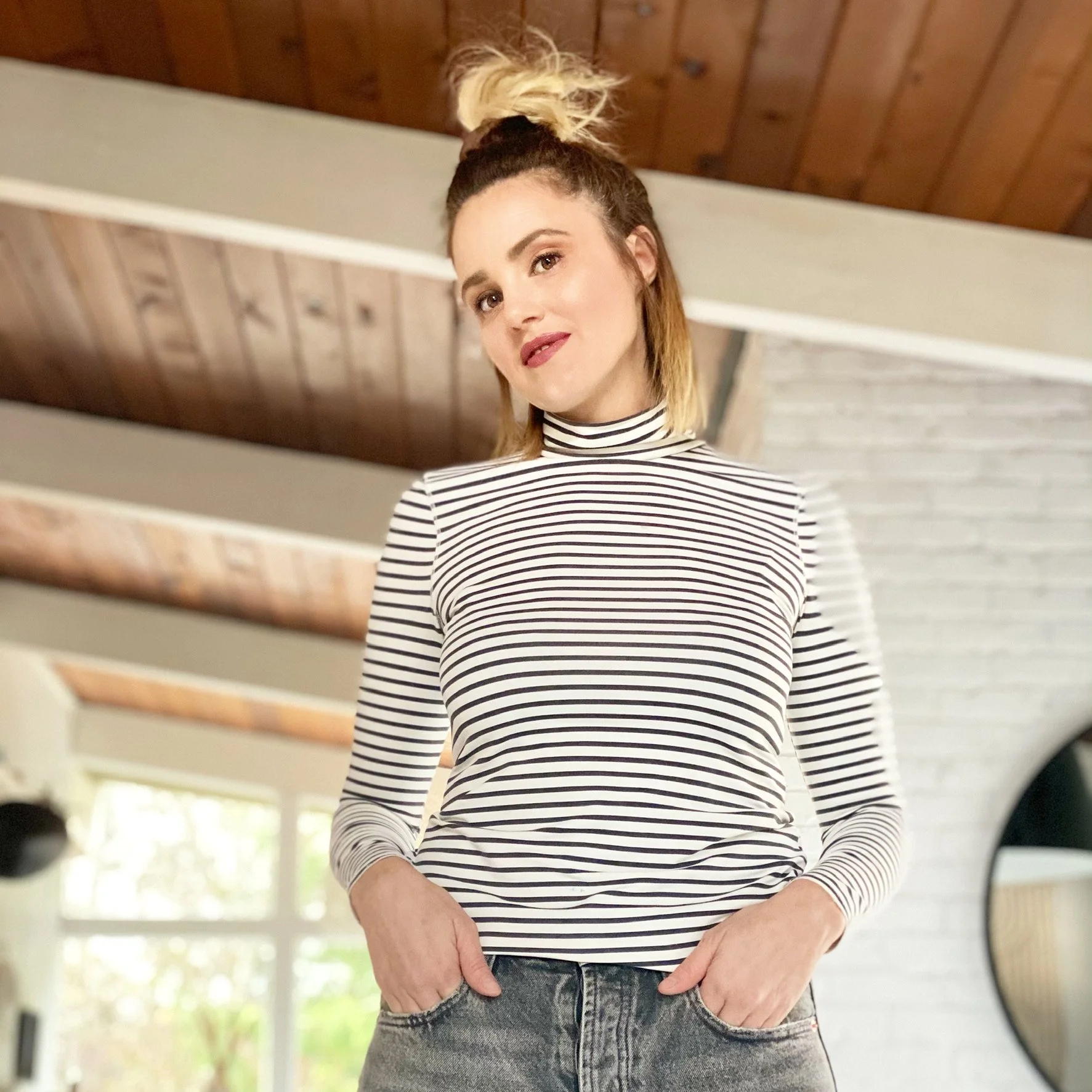 A woman with blonde hair tied in a messy bun wearing a black and white striped long-sleeve shirt and distressed jeans, standing inside a house with wooden ceiling and large windows showing greenery outside.