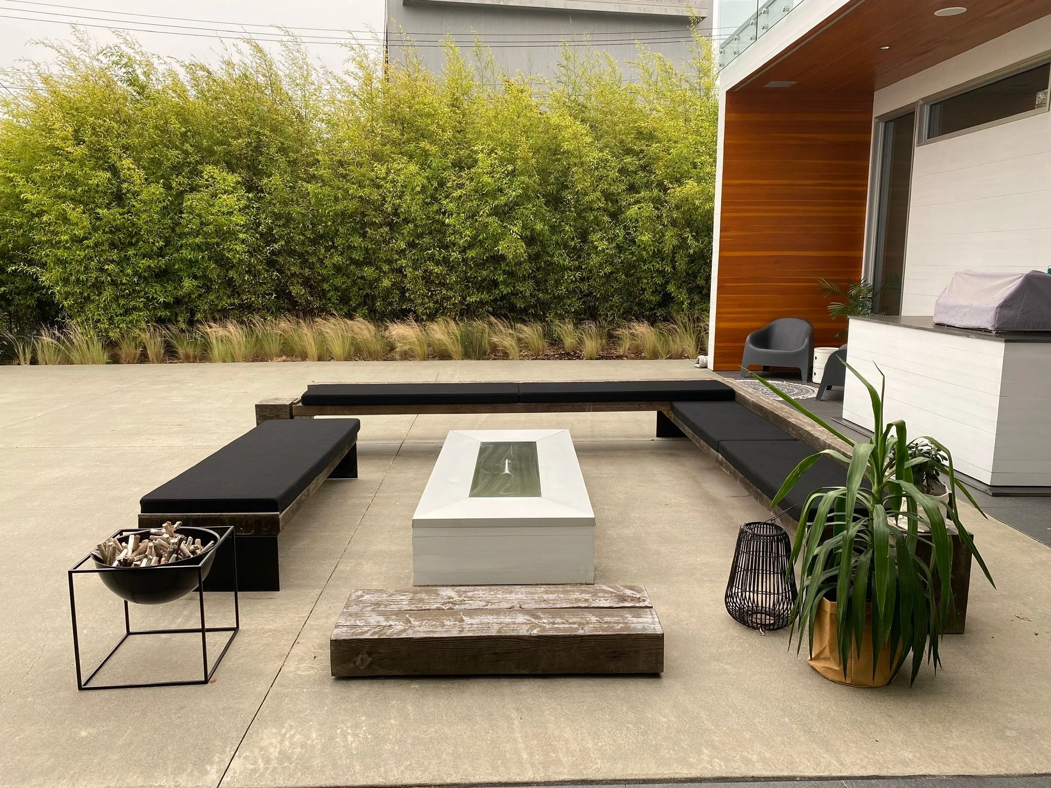 Outdoor patio area with seating and plants, including a large green shrub, a black chair, a white and wooden coffee table, and a potted plant with long leaves. Modern house with white walls, wooden paneling, and large windows in the background.