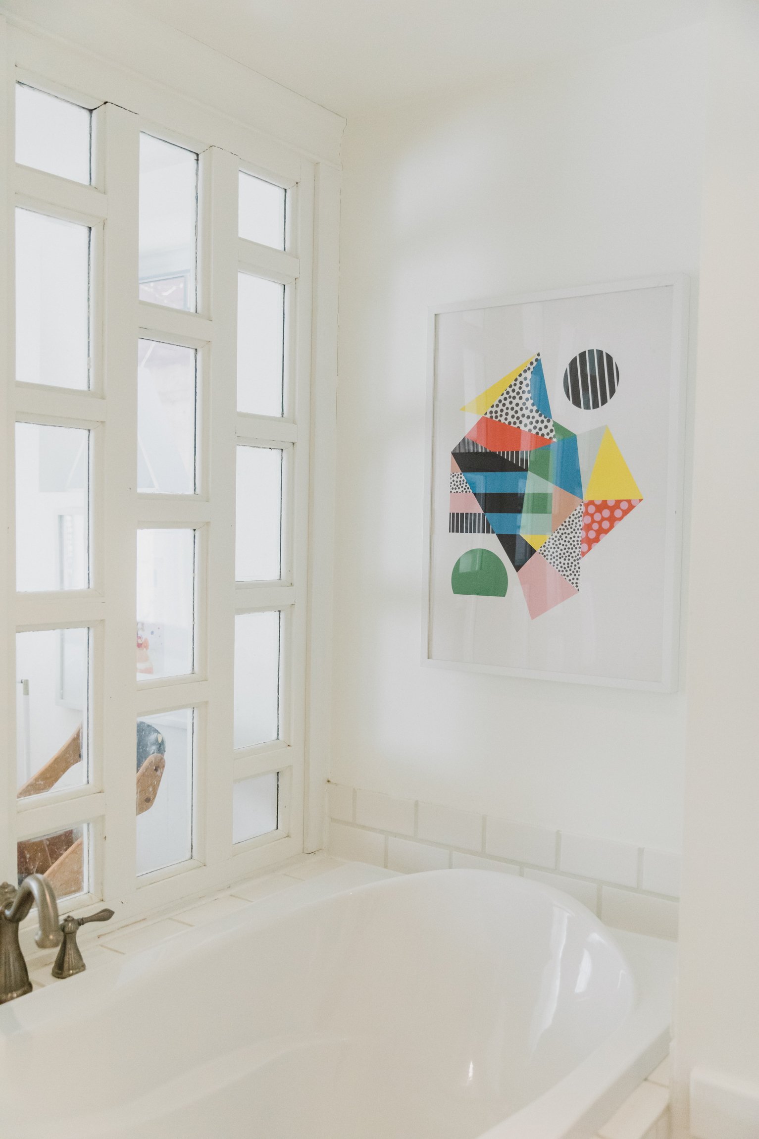 White bathtub with a metal faucet in a bathroom with a window and colorful abstract artwork on the wall.