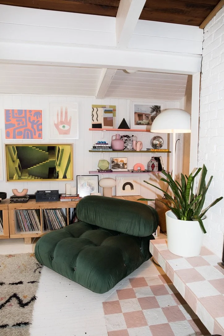 Interior of a modern living room with a green lounge chair, a wooden shelf with vinyl records, decorative art on the wall, and a large potted plant on a checkered pink and white side table.