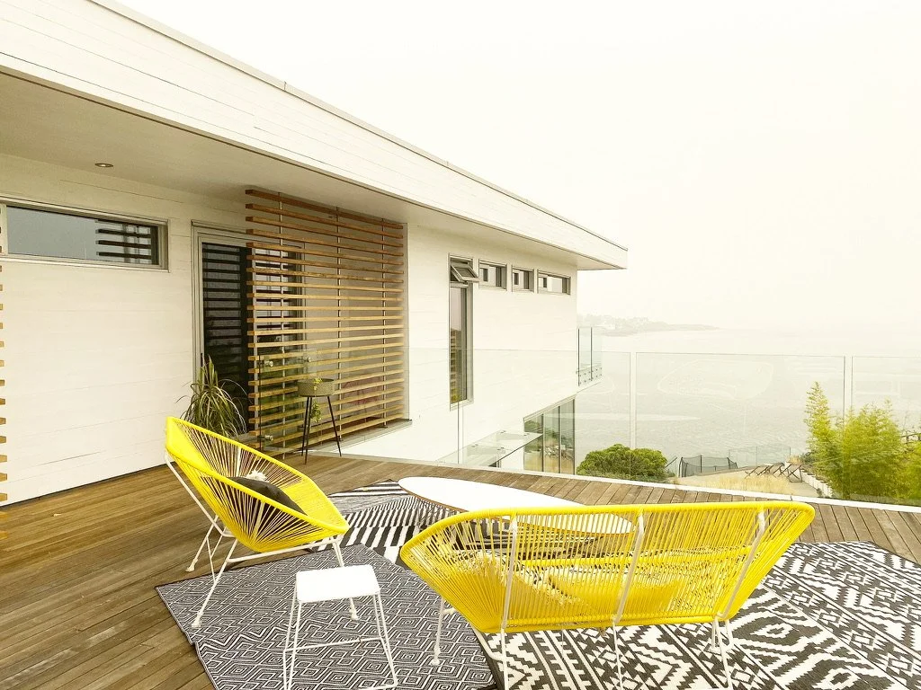 Outdoor balcony with yellow modern lounge chairs, a black and white geometric rug, glass railing, and view of a foggy or cloudy landscape.