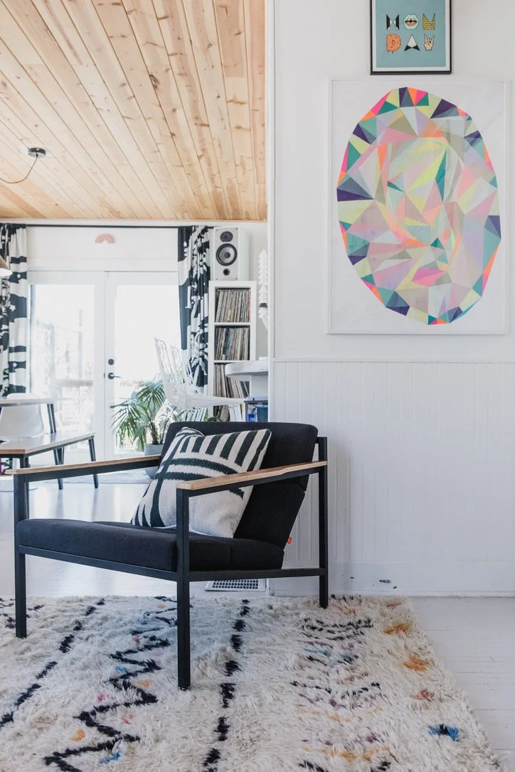 Living room with black armchair on a patterned rug, wall art with colorful geometric portrait, white walls, wooden ceiling, and glass double doors with black and white curtains. There is a small bookshelf and a plant near the doors.