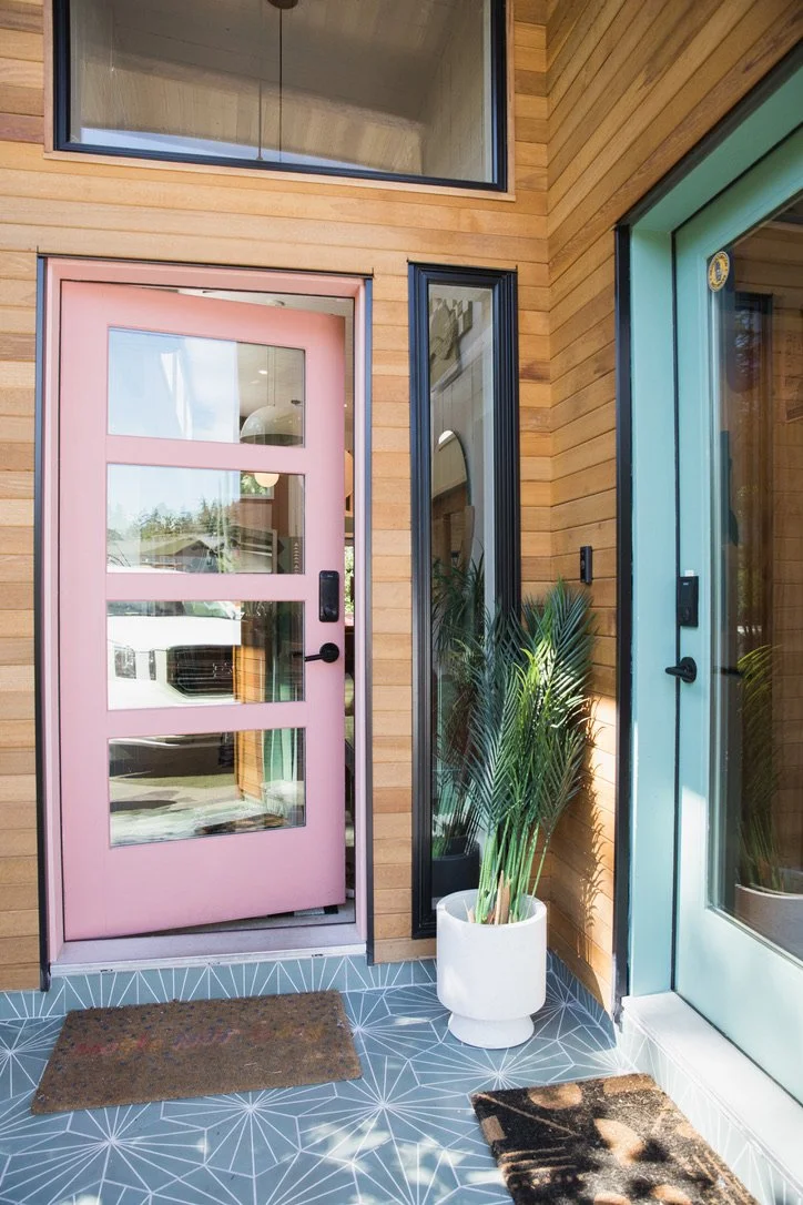 Exterior view of a house featuring a pink front door with glass panels, a large window above, a white potted plant, and a glass side door, all with wooden siding and geometric-patterned tiles on the porch.