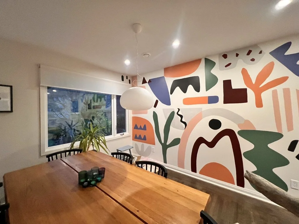 Modern dining room with a wooden table, black chairs, a white pendant light, a window with a white blind, a tall plant near the window, and an abstract mural with various shapes and colors on one wall.