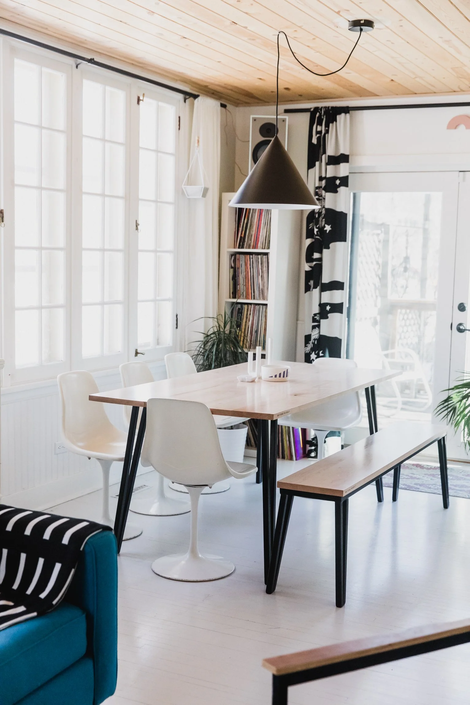 Interior of a bright dining area with a light wood ceiling, large windows with white curtains, a black and white patterned curtain, a modern black and wood dining table with white chairs, a blue armchair with striped clothing, a hanging black conical pendant light, a bookcase with vinyl records, and a sliding glass door leading outside.