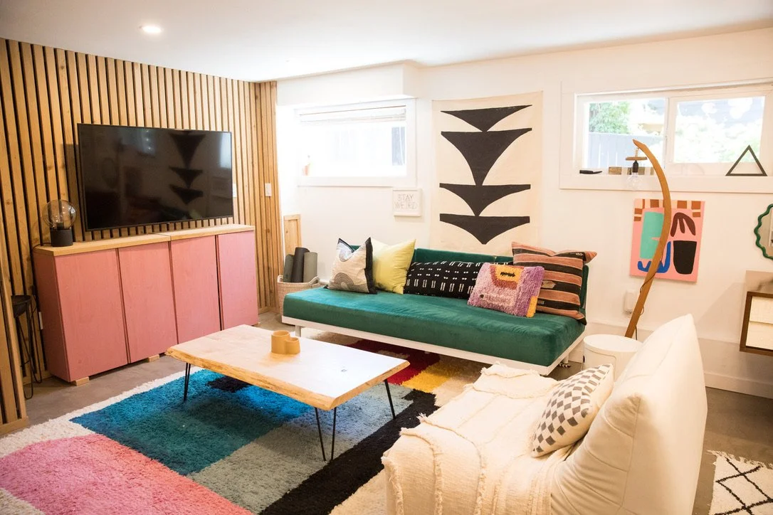 Living room with a wall-mounted flat-screen TV, pink cabinet, colorful rug, white couch, green sofa, and art decor on white walls and windows.