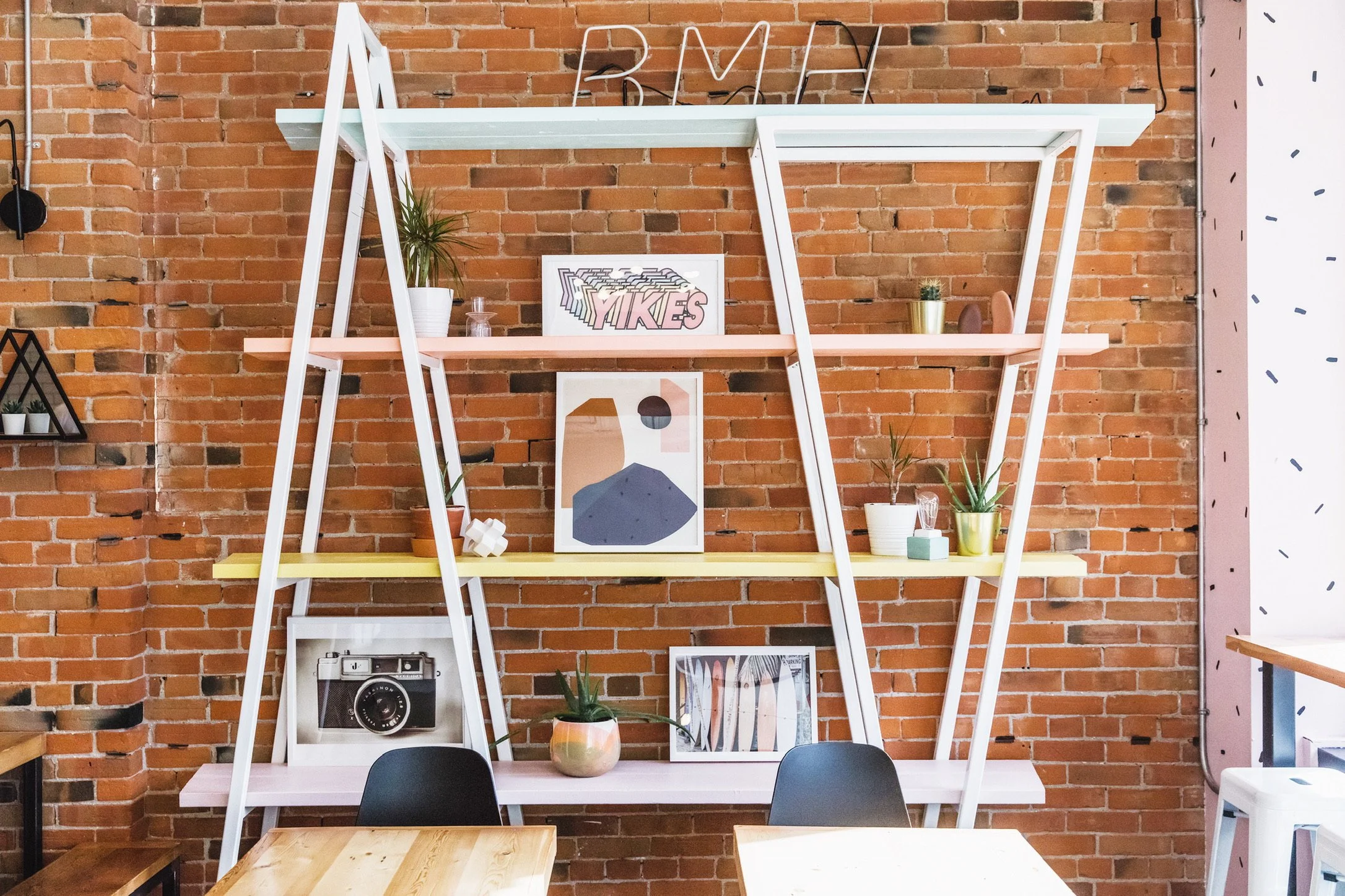 A decorative interior wall with exposed brick, featuring a white shelf with colorful accents and decor including framed artwork, potted plants, and vintage camera.