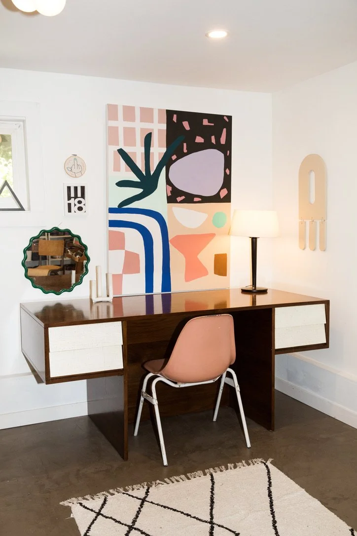 Interior of a home office with a wooden desk, pink chair, modern art on the wall, a lamp, and decorative wall pieces, with a window and a mirror reflected in the small mirror on the wall.
