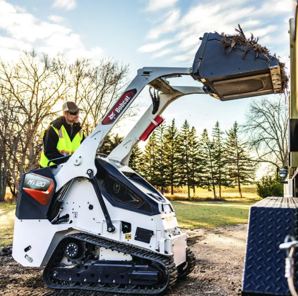 A person wearing a yellow safety vest and a hat operating a Bobcat mini track loader with a front bucket attachment filled with dirt and sticks, outdoors on a dirt path with trees and a blue sky in the background.