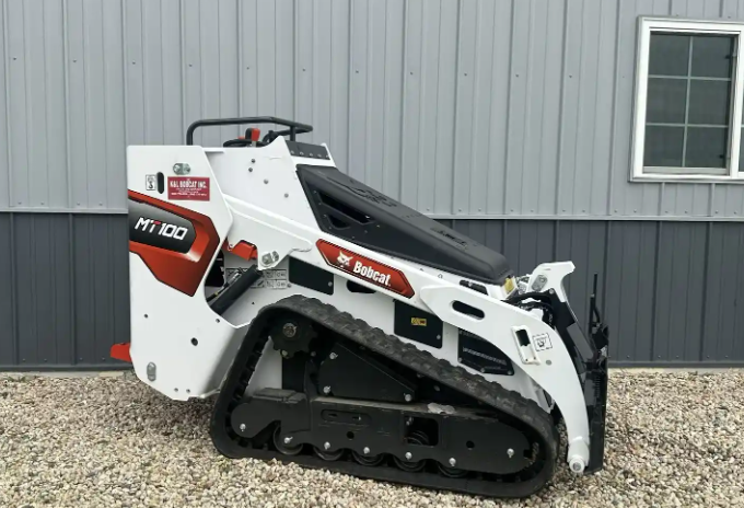 A Bobcat MT100 compact track loader parked outdoors on gravel in front of a metal-sided building with a window.