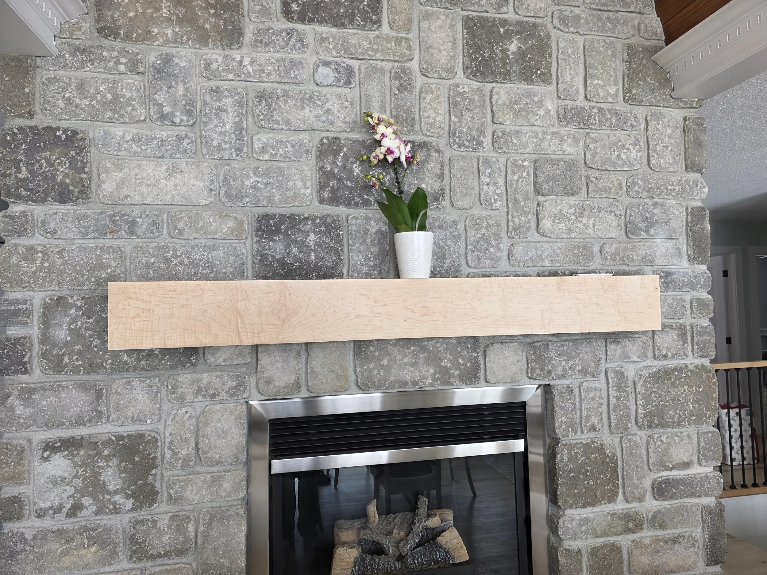 A brick fireplace with a wooden mantel, decorated with a white flower pot containing an orchid. The bricks are gray and weathered, and there is a black fireplace below the mantel with stacked logs inside.