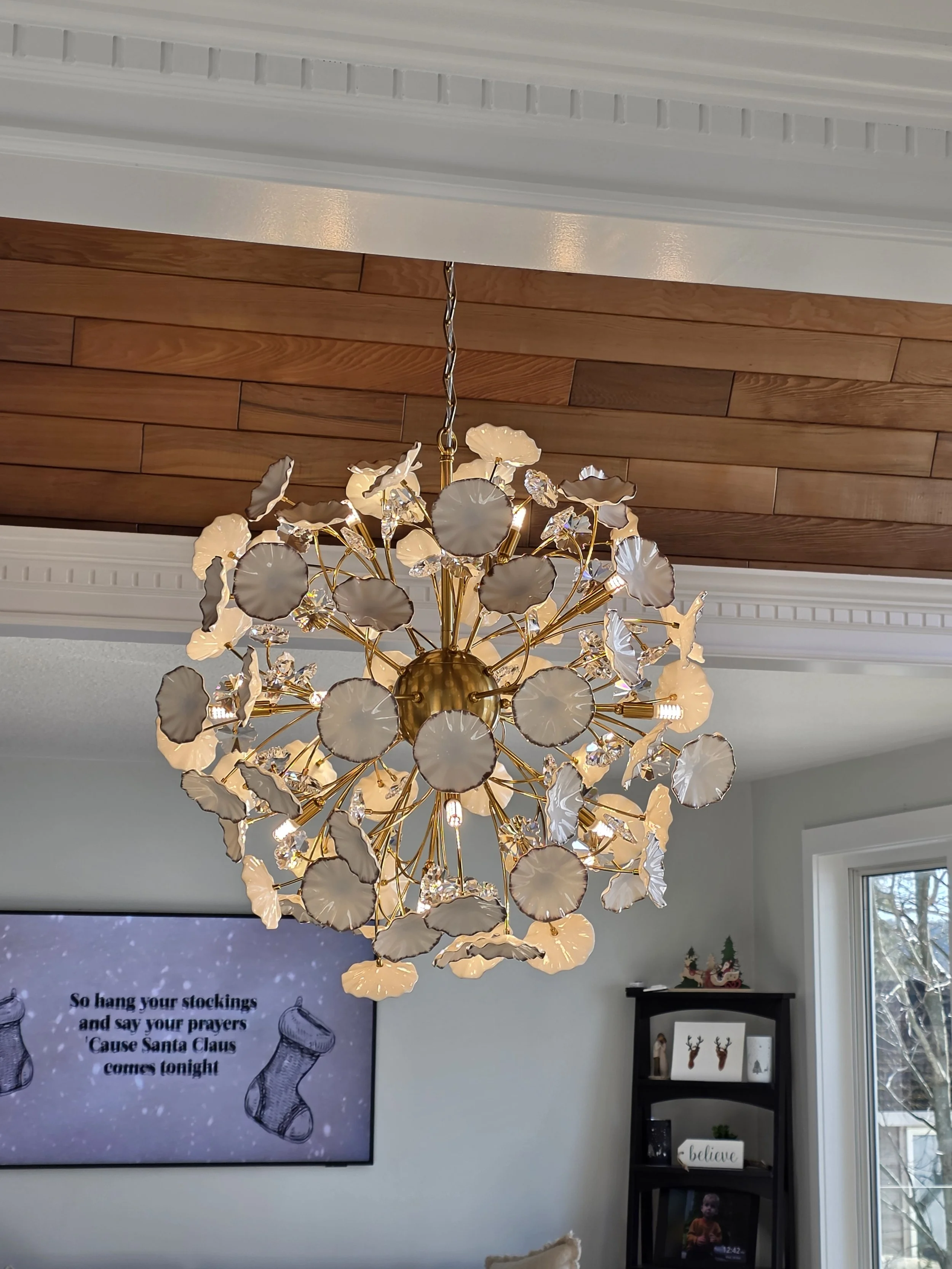 A gold chandelier with white and clear glass floral-shaped accents hanging from a white ceiling with crown molding and a wooden panel.