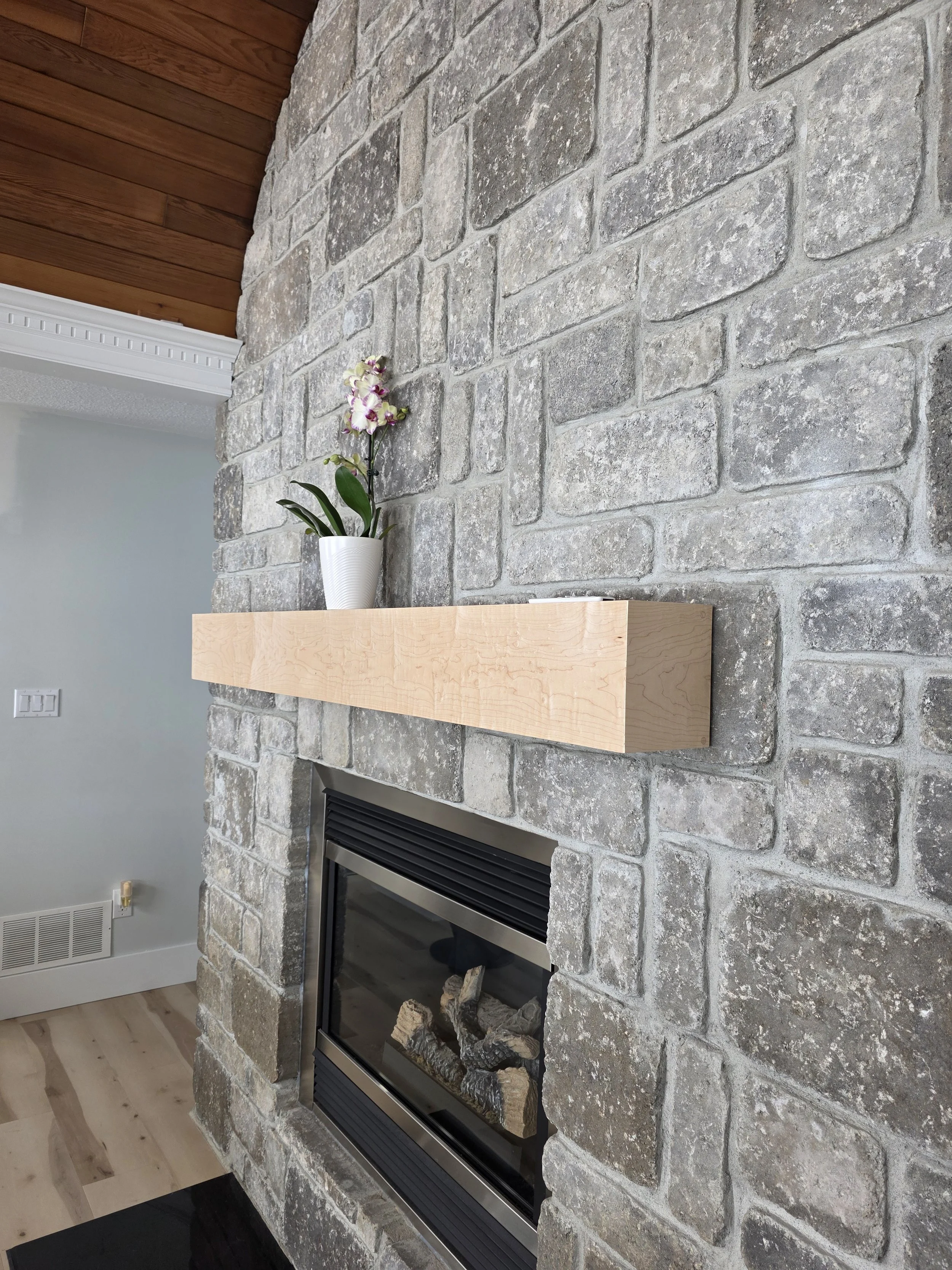 A stone fireplace with a concrete mantle, decorated with a white vase holding purple and white orchids, in a room with wooden ceiling and hardwood flooring.
