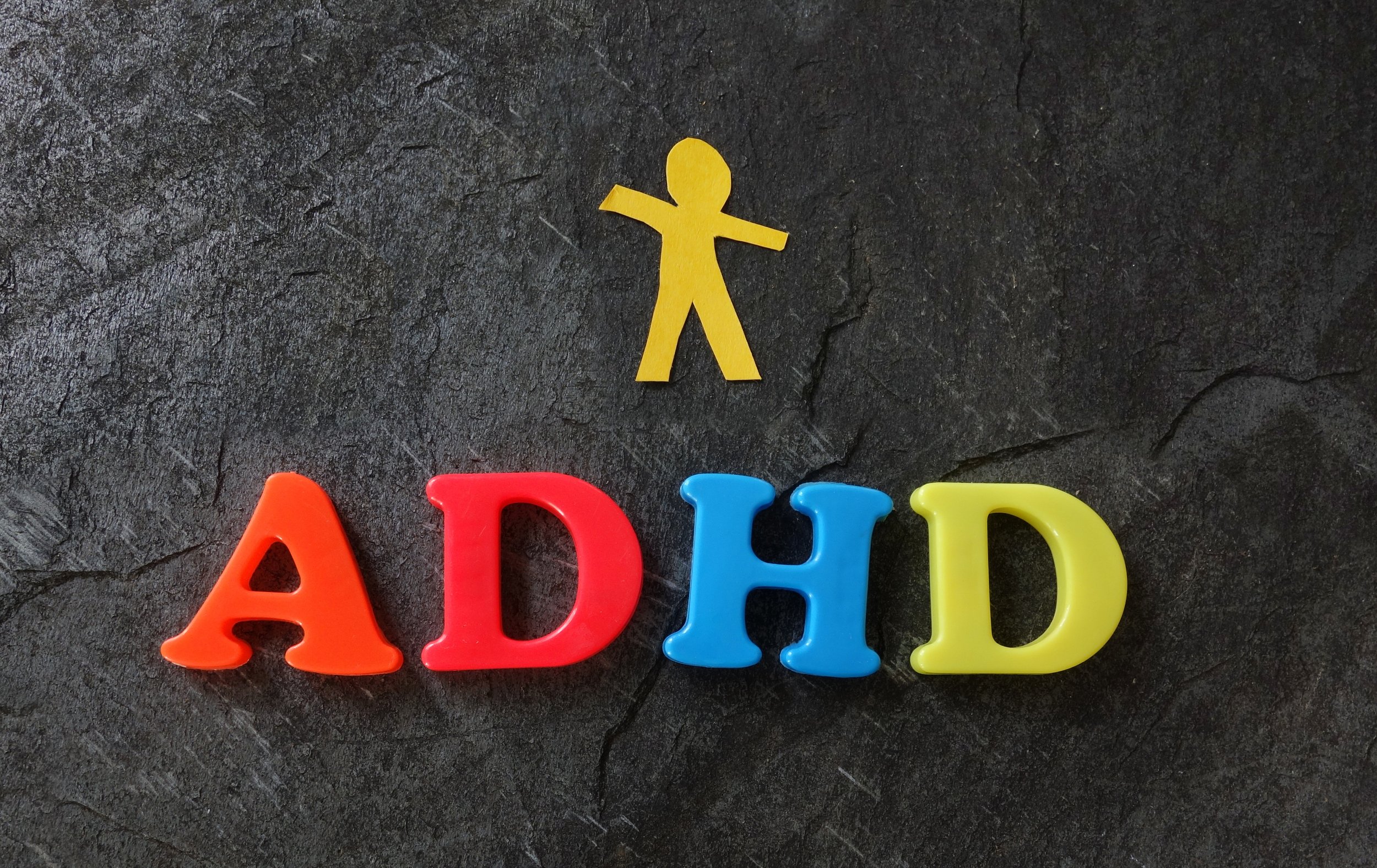 Colorful plastic letters spelling 'ADHD' with a yellow paper cutout of a person above them, on a dark textured surface.