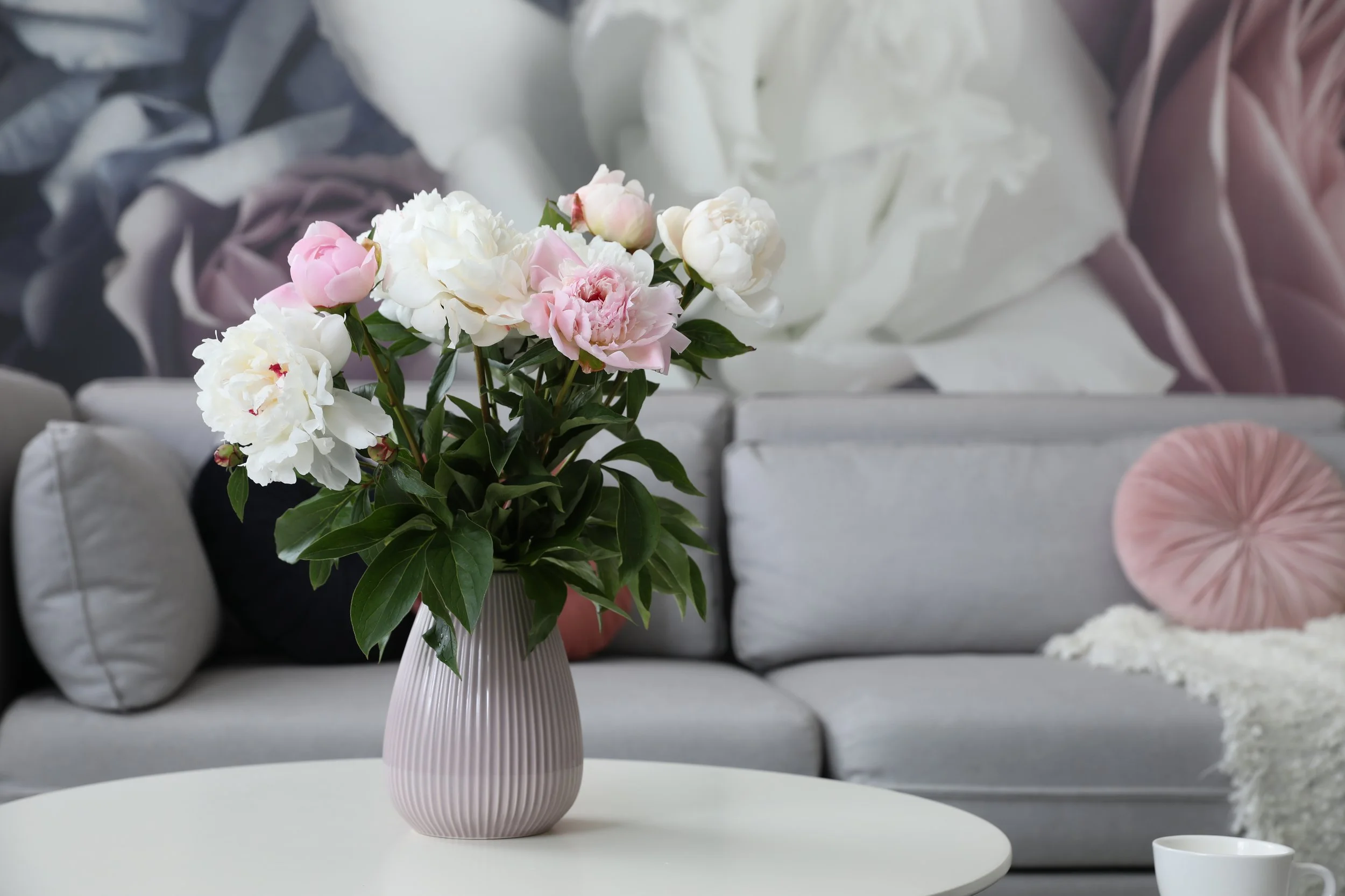 A vase with pink and white peonies on a white table in front of a gray sofa with pillows and a round pink throw pillow, with a decorative blanket or fabric in soft pastel tones in the background.