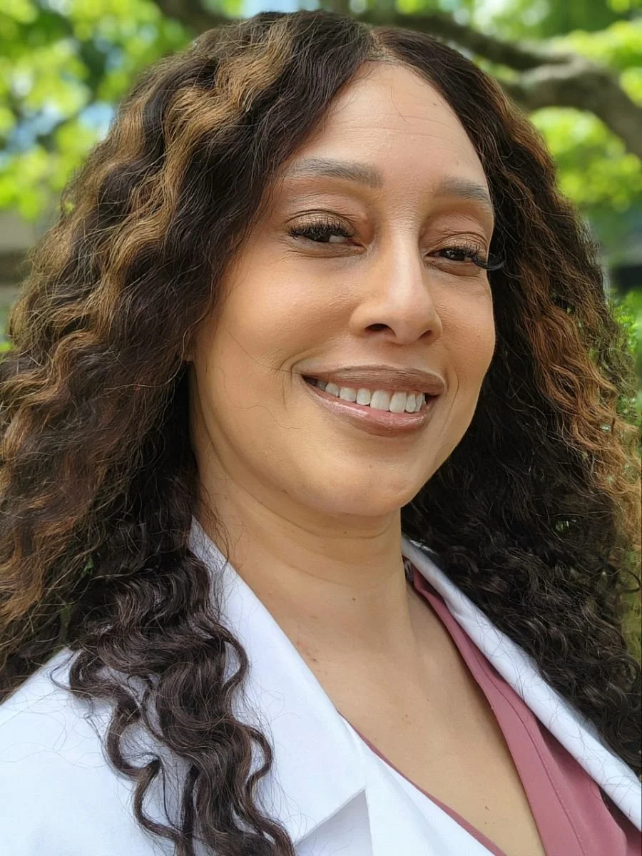 A woman with curly brown hair smiling outdoors with green leaves in the background.