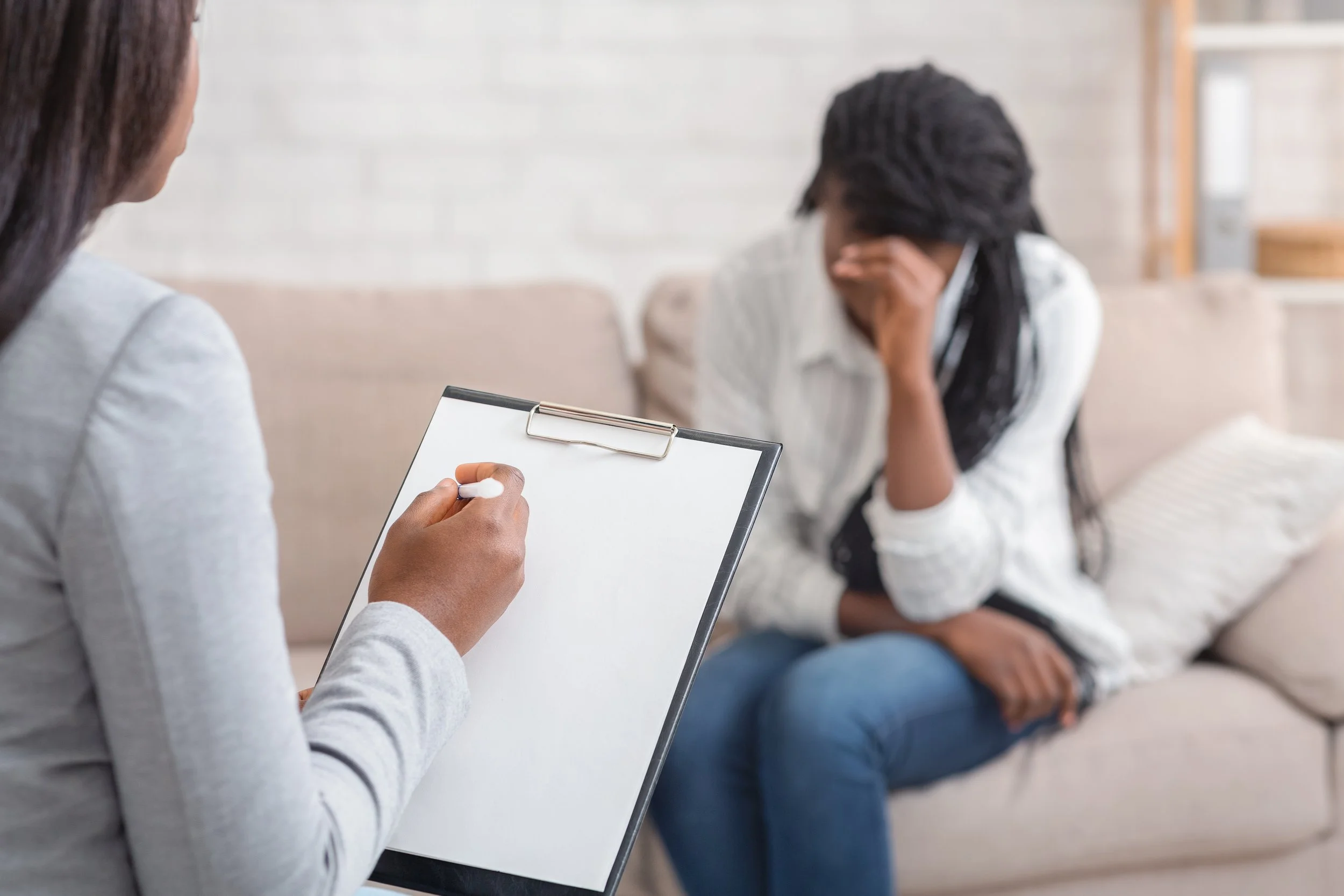 A woman sitting on a sofa, covering her face with her hand, during a therapy session with a counselor taking notes on a clipboard.