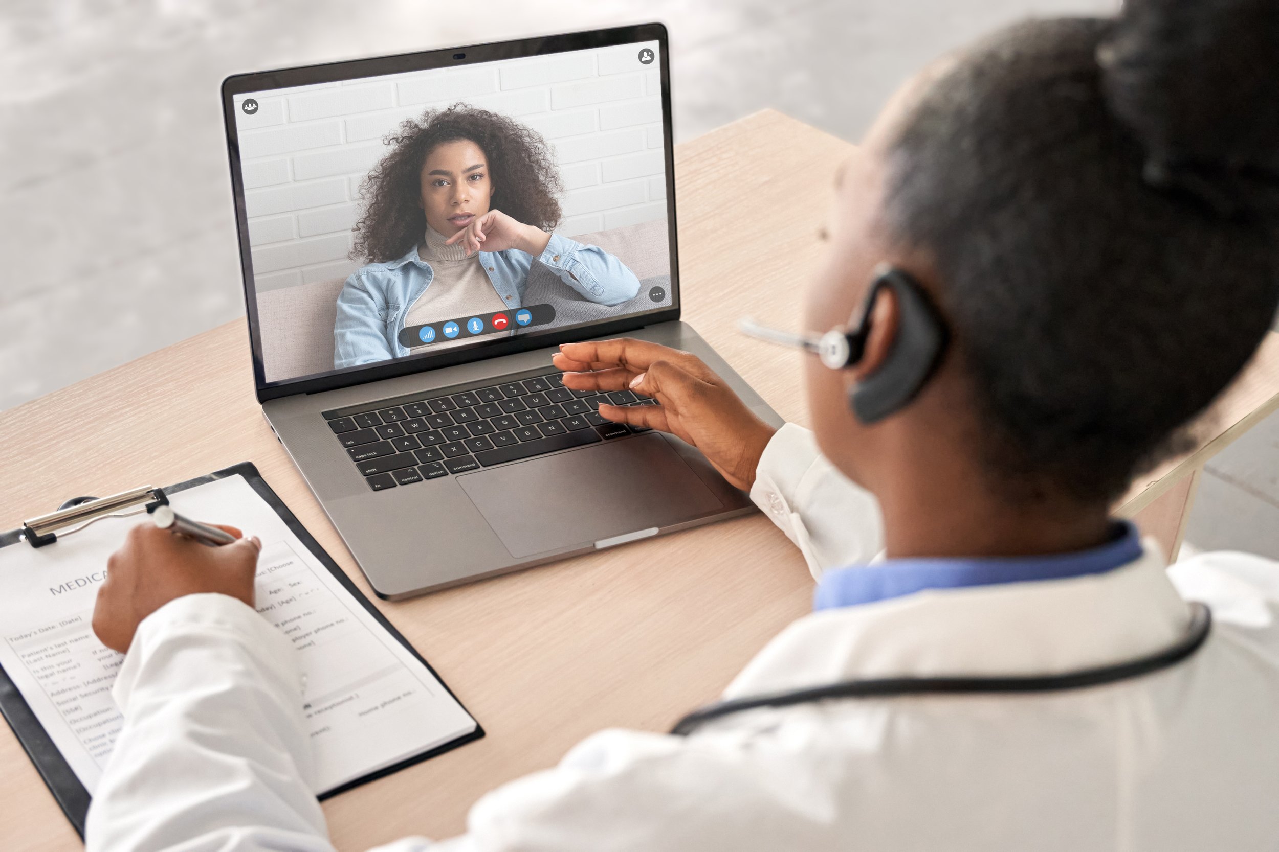 A doctor having a video consultation with a woman on a laptop at a desk. The doctor is wearing a white coat and a headset, taking notes on a clipboard.