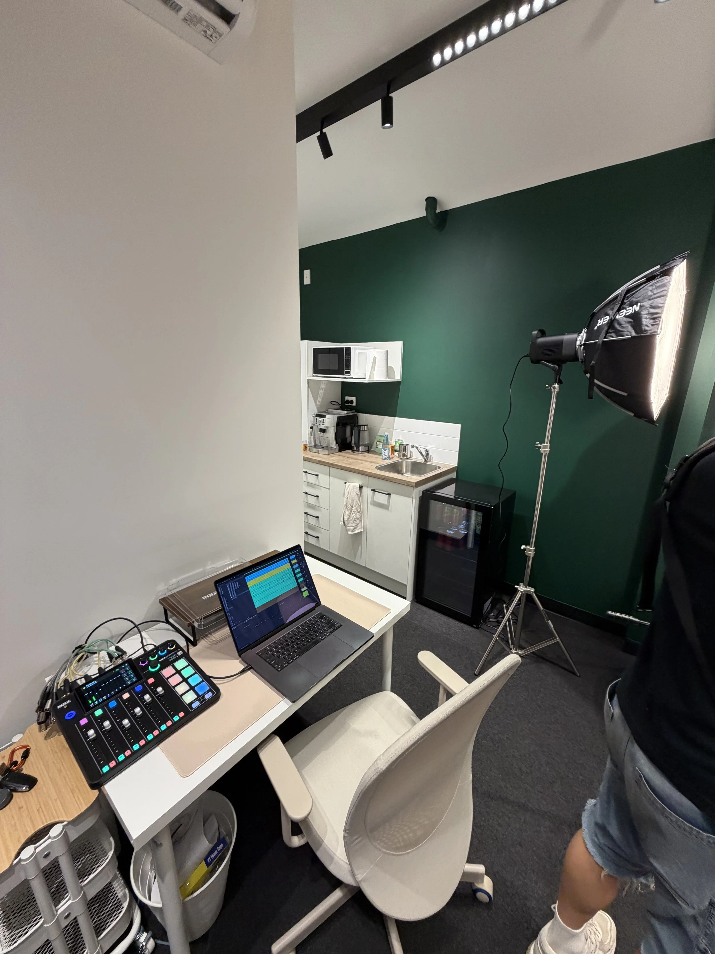 A room set up for video or photo production featuring a green screen wall, a studio light with a softbox, a computer on a desk with audio and visual equipment, a small kitchenette with microwave, coffee machine, and sink, a white chair at the desk, and a person wearing shorts and sneakers standing near the setup.