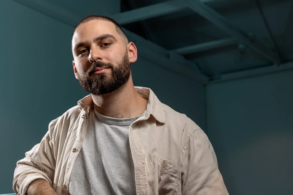 A man with a beard and short hair wearing a light beige shirt over a gray t-shirt, standing indoors against a teal wall.