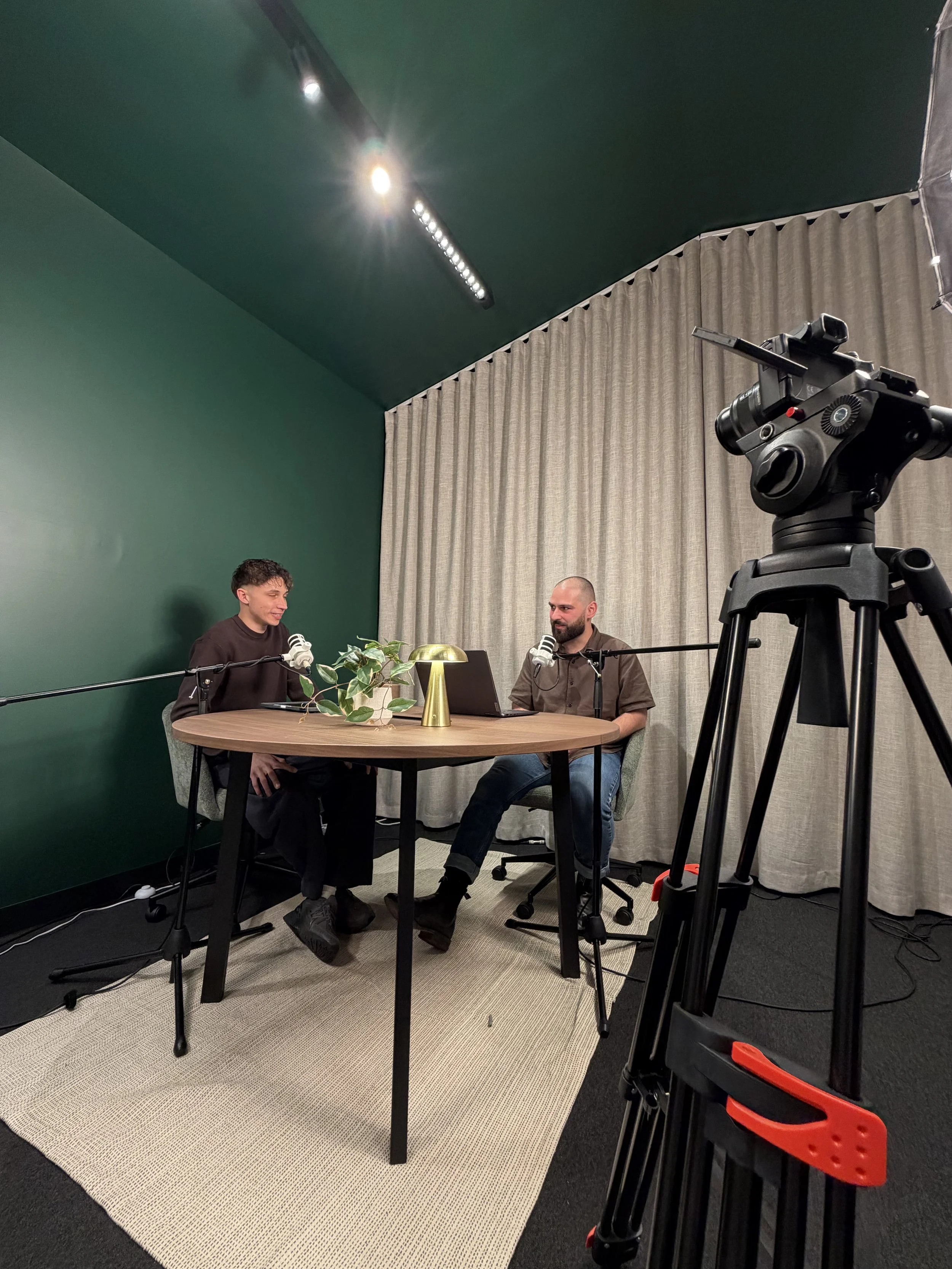 Two men sit at a round table in a room with a green wall and dark ceiling, with a large camera on a tripod recording them, microphones on the table, a laptop, a decorative plant, and a table lamp.