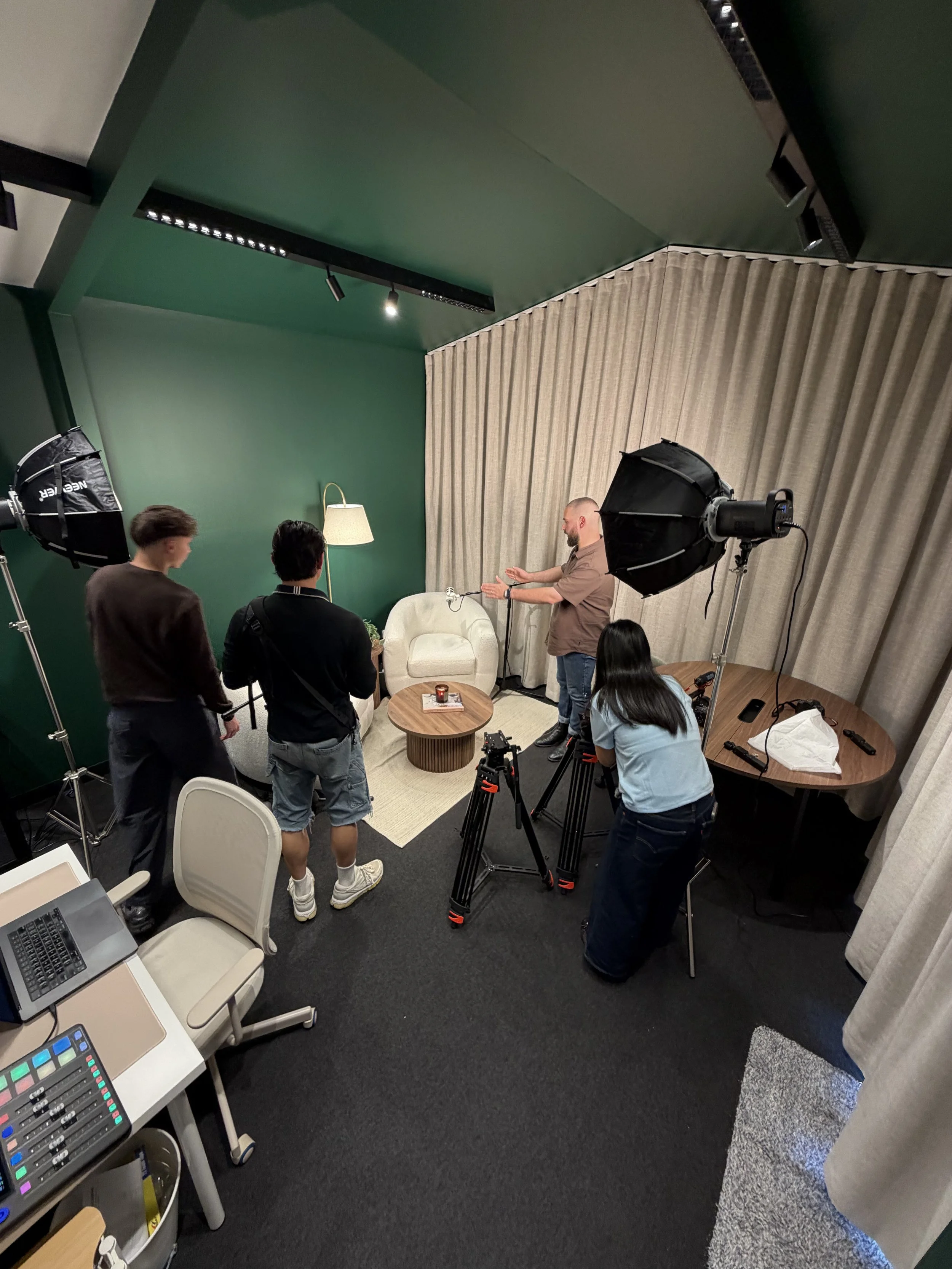 Setup for a video or photo shoot inside a green-ceiling room with four people preparing, including lighting equipment, a white chair, a small round table, and a laptop on a side table.
