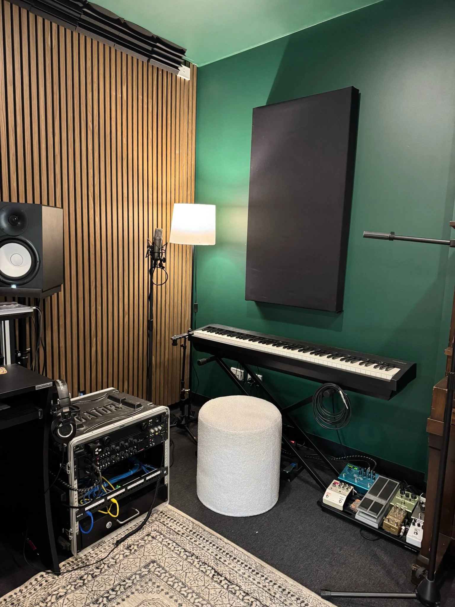 Music studio with green walls, wooden acoustic panel, electronic keyboard, sound equipment, microphone on stand, studio monitor speaker, and various effects pedals and audio gear.