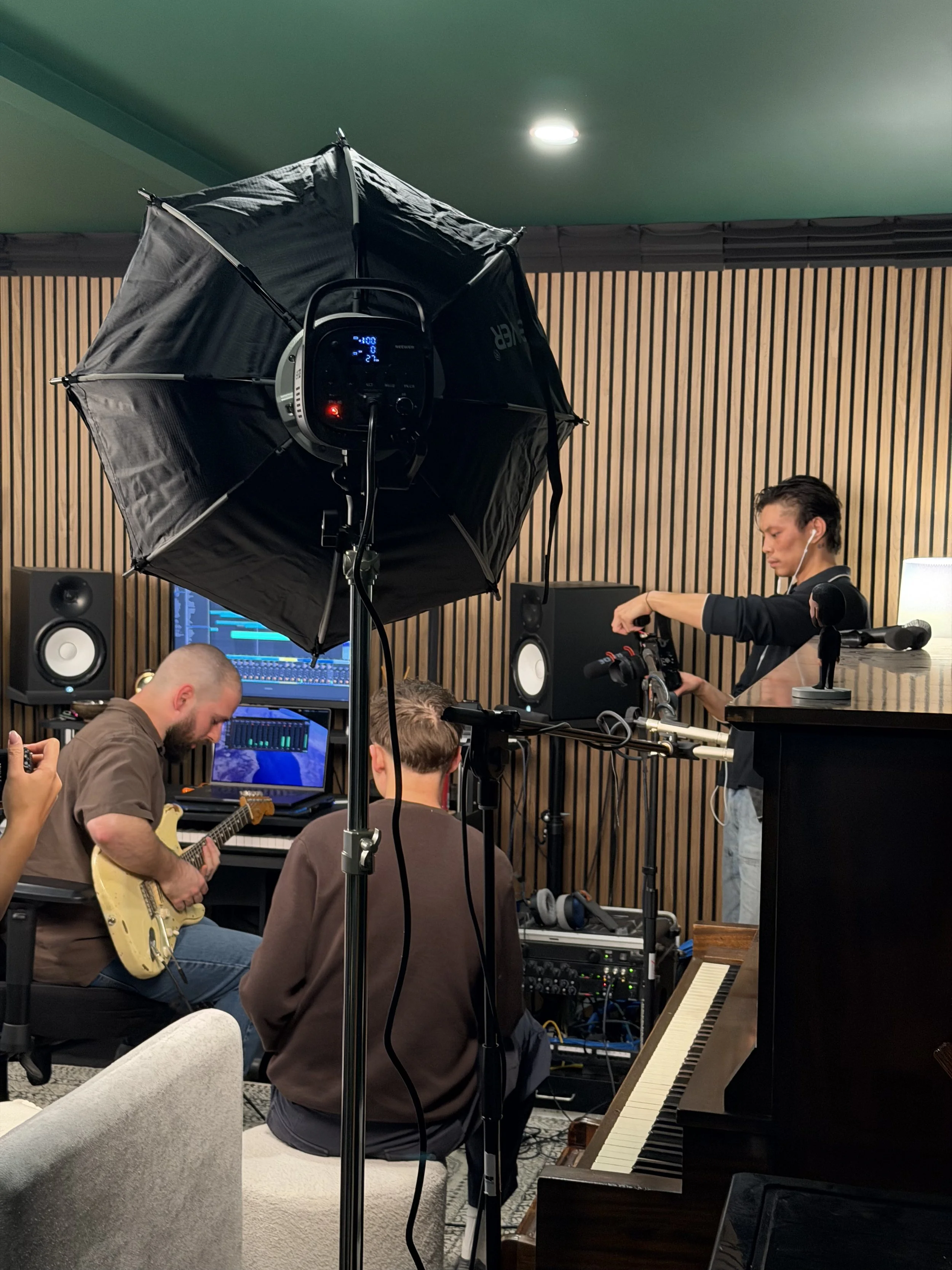 A recording studio with musicians playing instruments, including a guitarist and a piano, while a person adjusts equipment. A large studio light is set up in the foreground, and soundproof wooden panels line the wall.