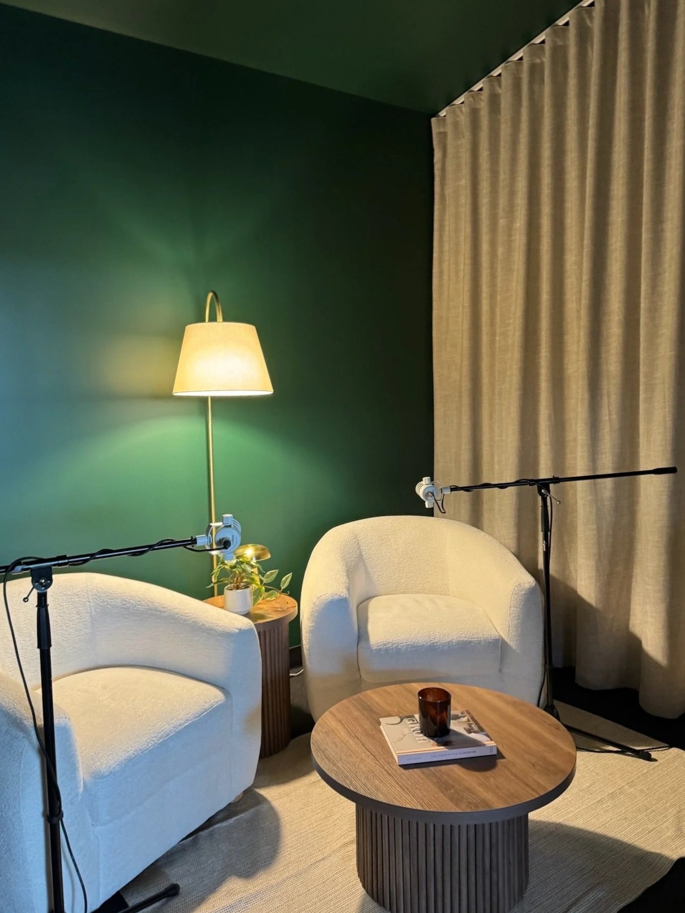 Comfortable indoor seating area with two white armchairs, a small wooden side table, a larger round wooden coffee table with a book and a dark brown cup, beige curtains, a green accent wall, a tall floor lamp, and microphone stands.