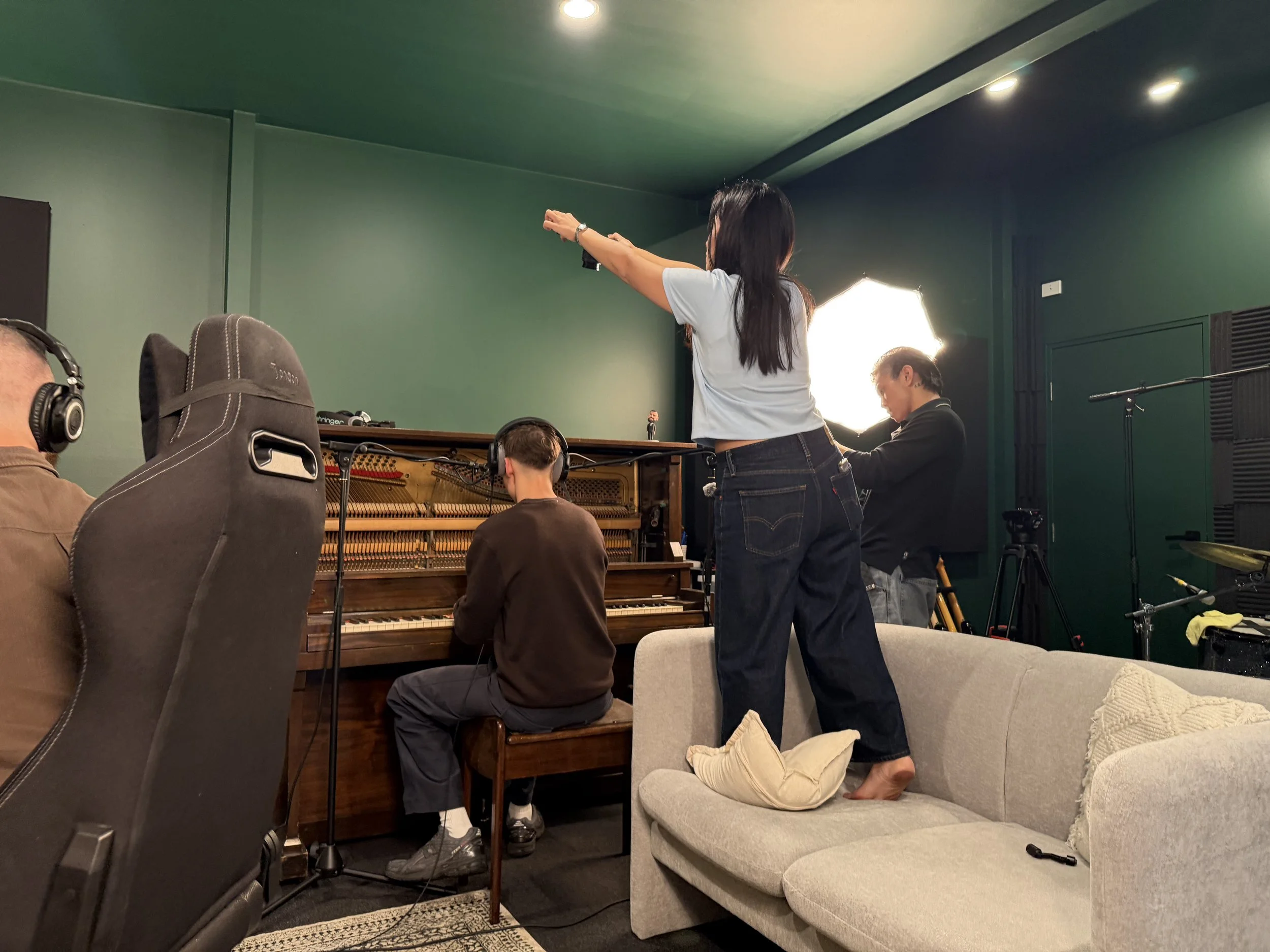 A woman with long dark hair and a white t-shirt stands on a sofa with her arms extended, pointing forward. She is in a room with green walls, a piano, and several people, some wearing headphones, are gathered around the piano. A man with gray hair and a black jacket is sitting at the piano. There is professional lighting and recording equipment in the room, indicating a music or recording session.