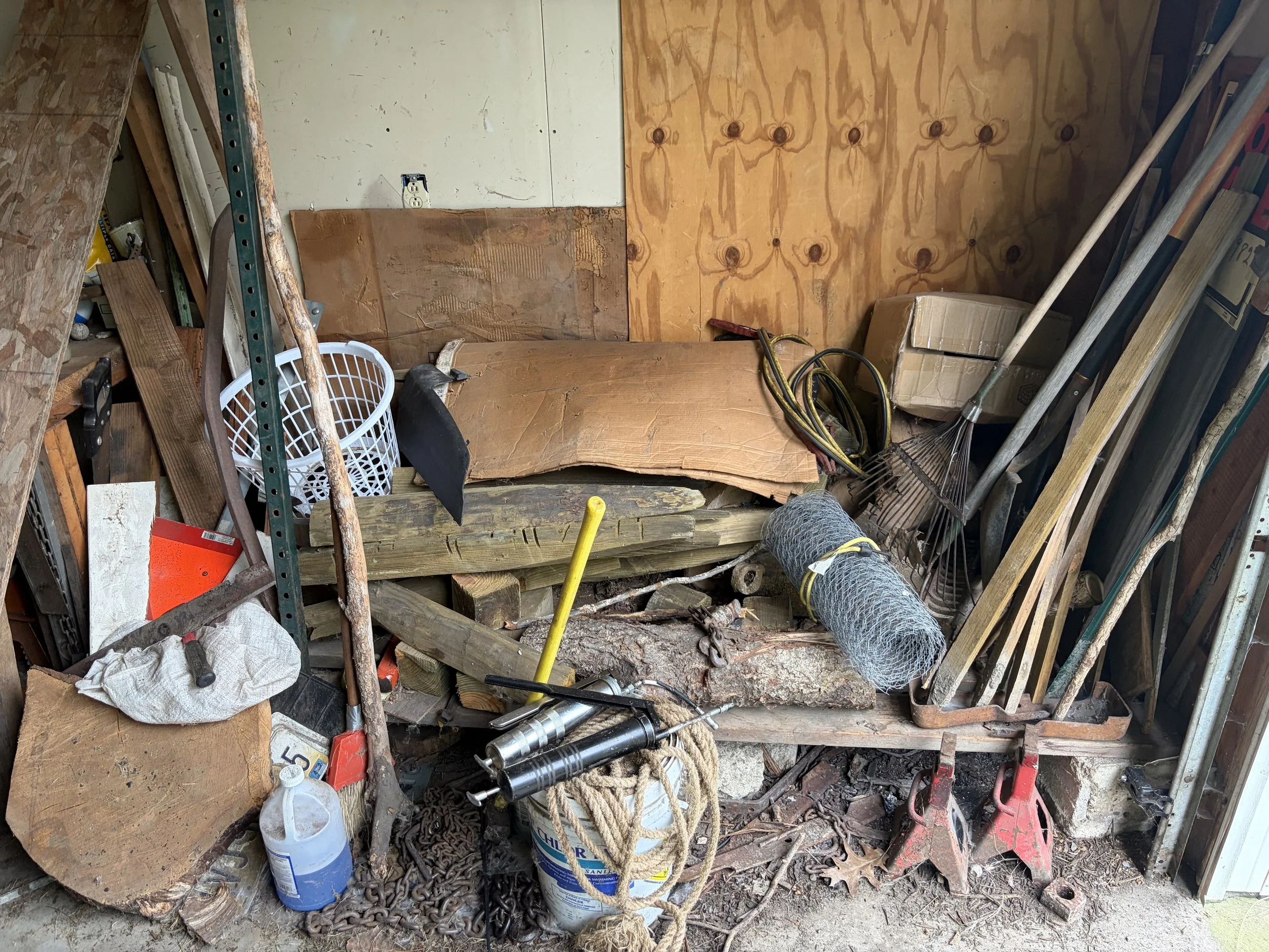 A cluttered storage space or garage with various tools, wood pieces, ropes, and containers scattered around. There is a wooden plank leaning against the wall, a black pipe with yellow and black cords, rakes, and a cardboard box, with a plywood wall in the background.