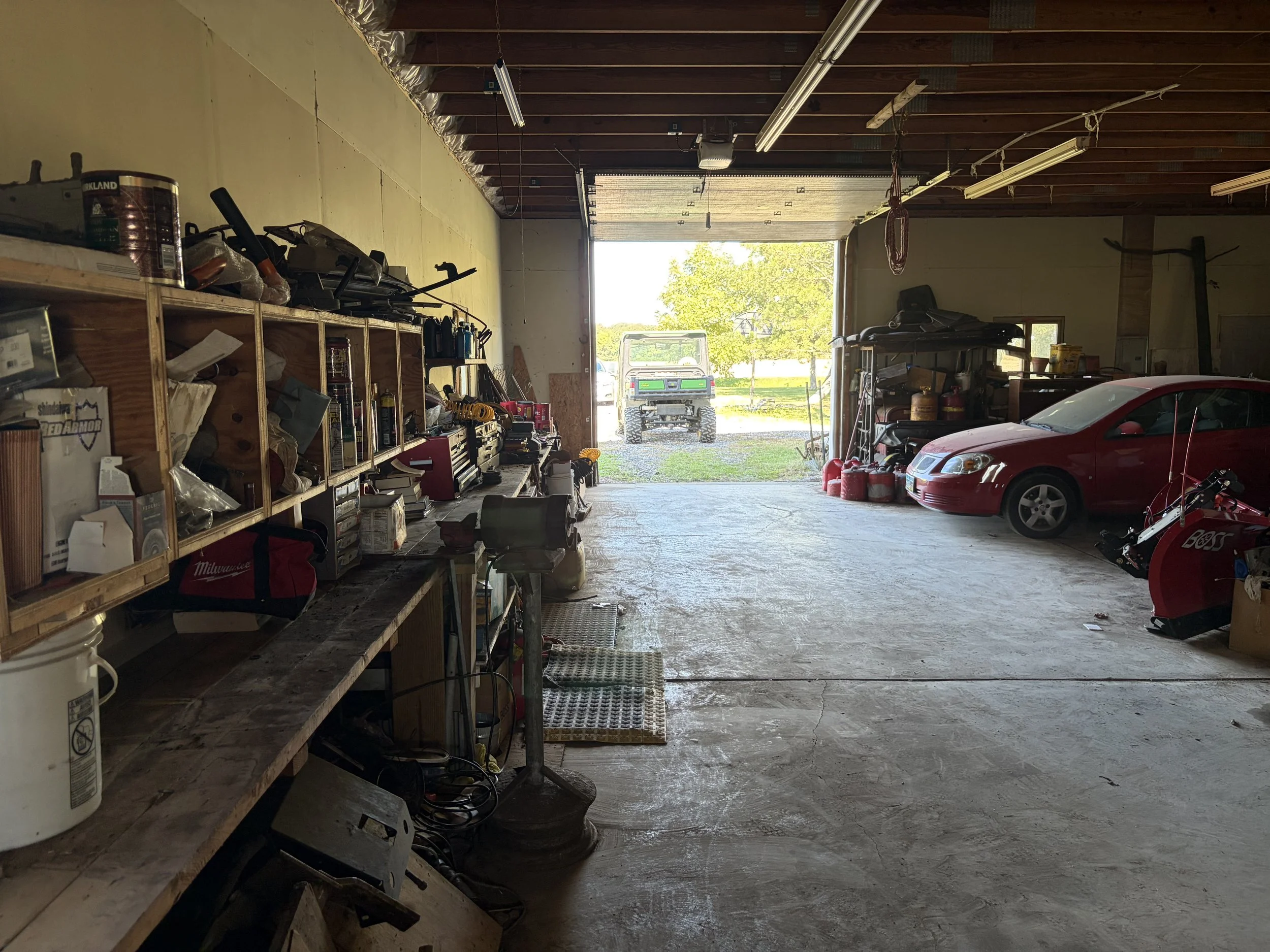 Inside a garage with shelves on the left filled with various tools and supplies, a red car parked on the right side, and an open garage door showing a lawn and a utility vehicle outside.