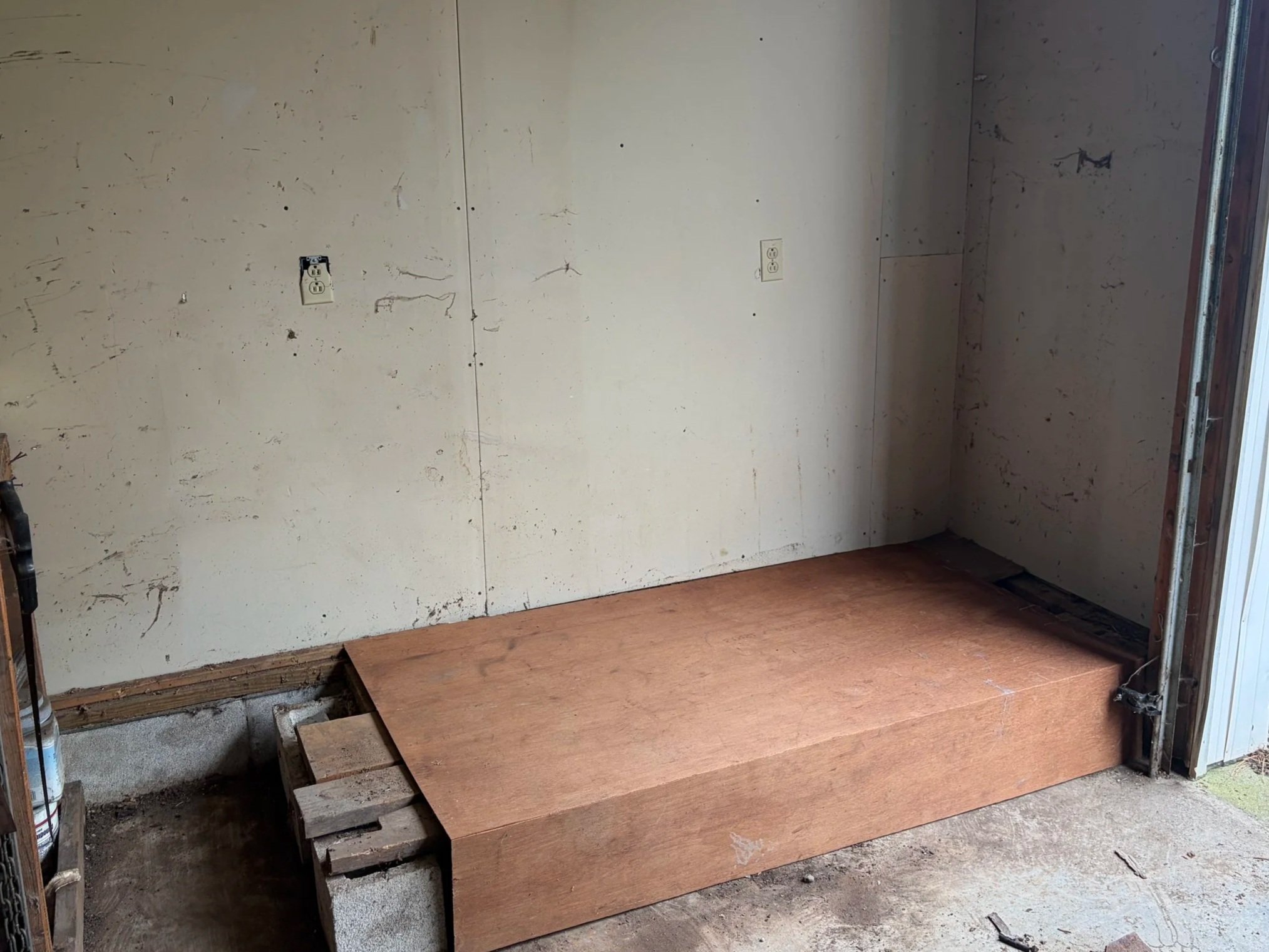 An unfinished room with a wooden platform, exposed support beams, and electrical outlets on the wall. The wall has marks and holes, indicating ongoing construction or renovation.