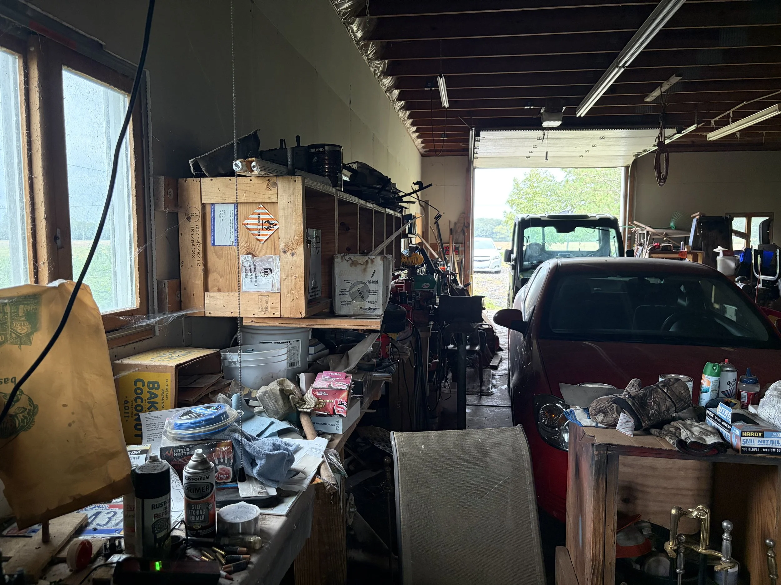 A cluttered garage with various tools, supplies, and a red car parked inside. The garage has a large open doorway, and shelves filled with miscellaneous items.