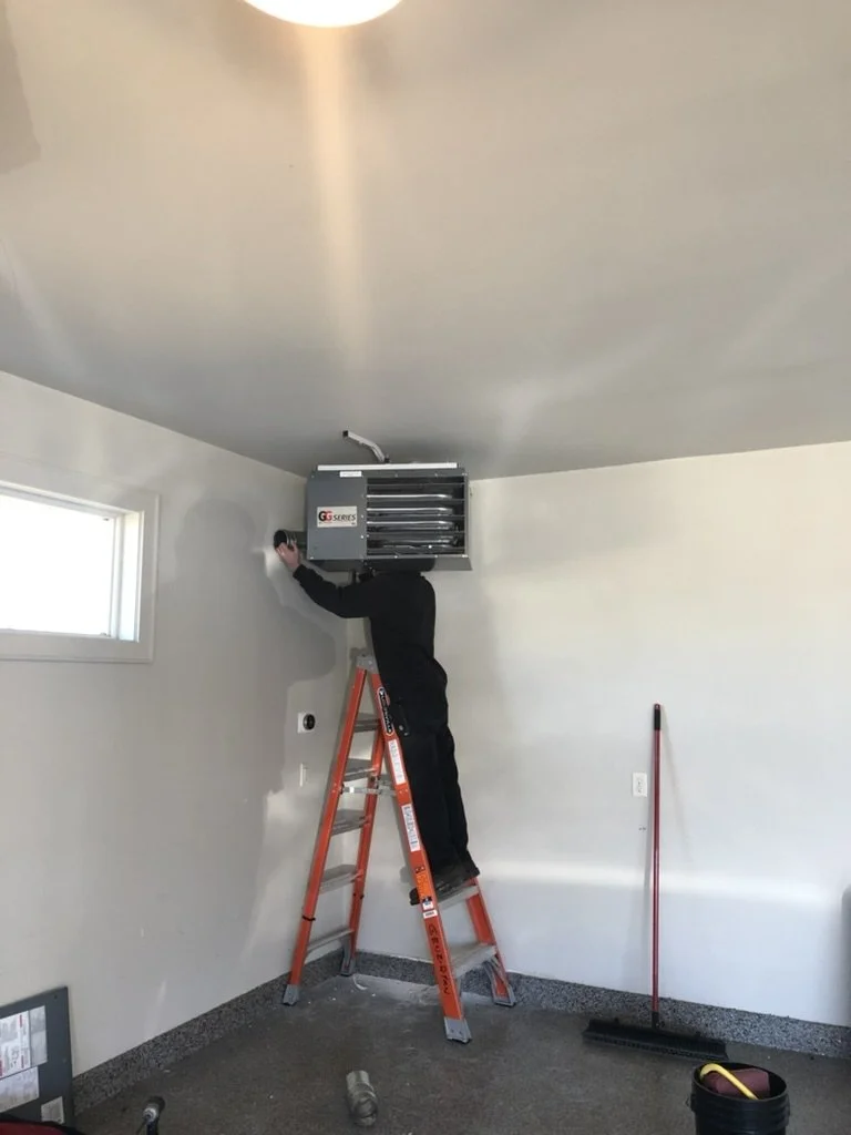 A person standing on an orange ladder installing or fixing an air conditioning unit in a room with gray walls and a window.