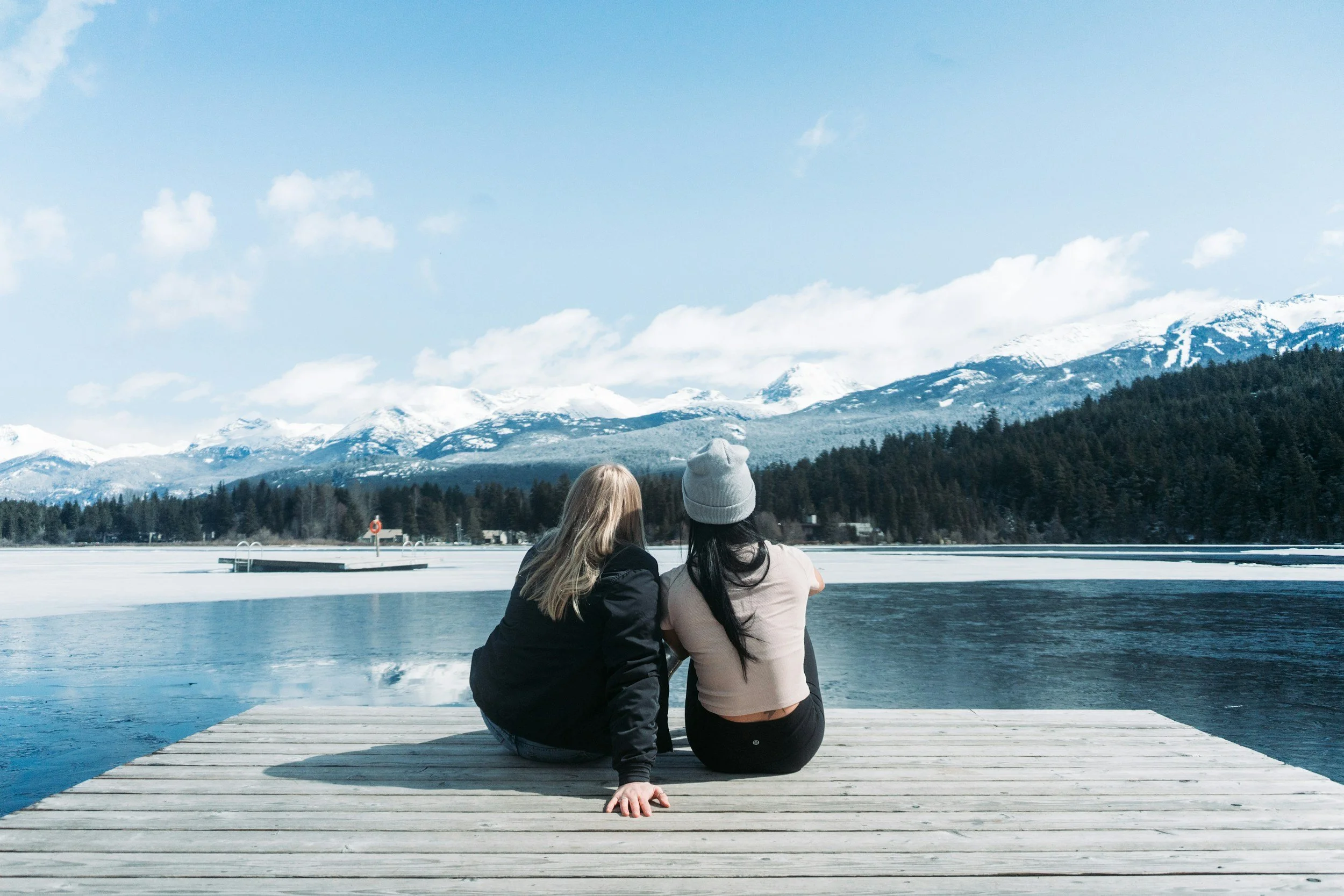 Two women sitting on a wooden dock overlooking a mountain lake with snow-covered peaks and evergreen forests