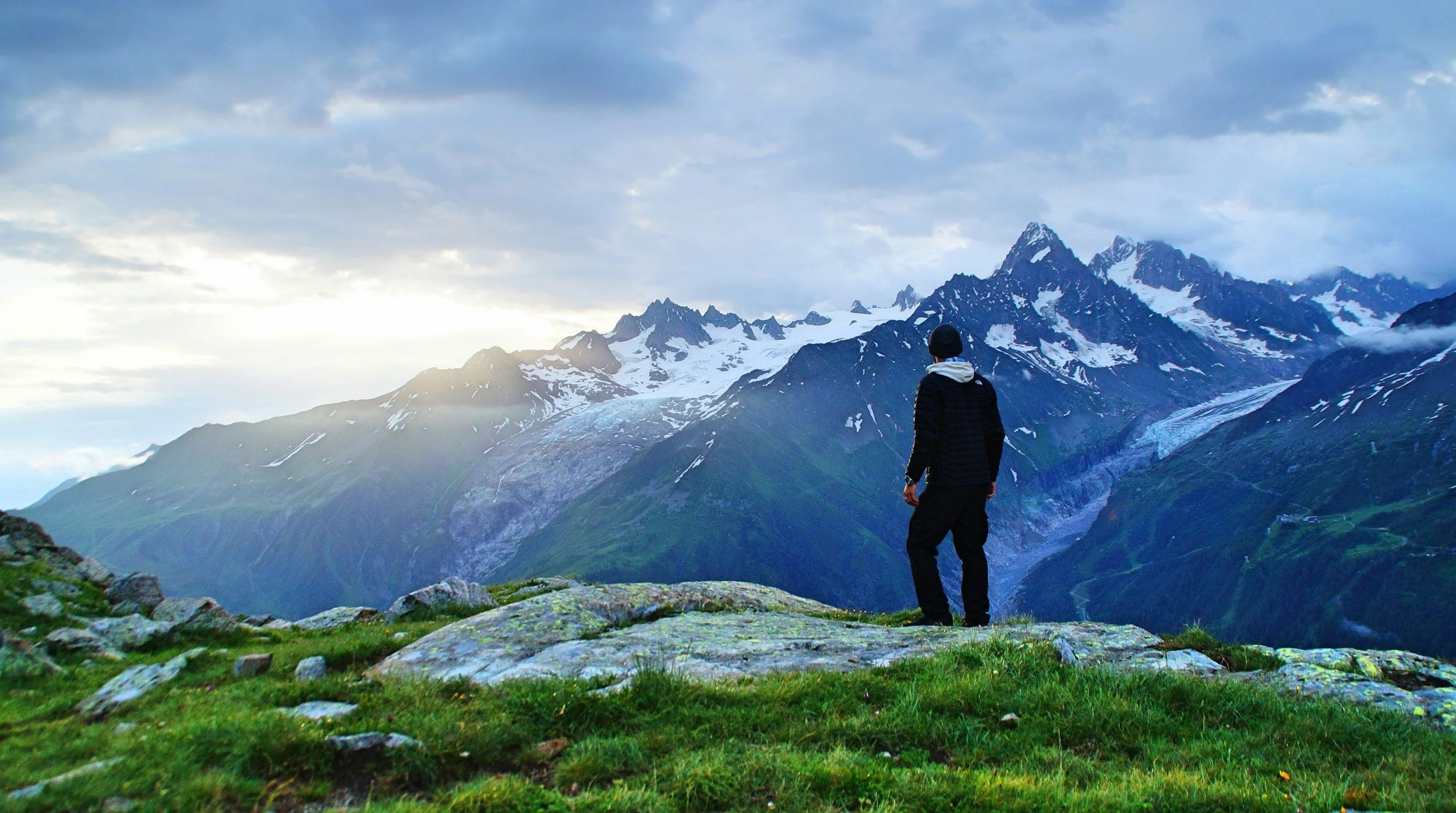 Hiker standing on a rocky alpine ridge overlooking snow-covered mountains and glaciers at sunrise