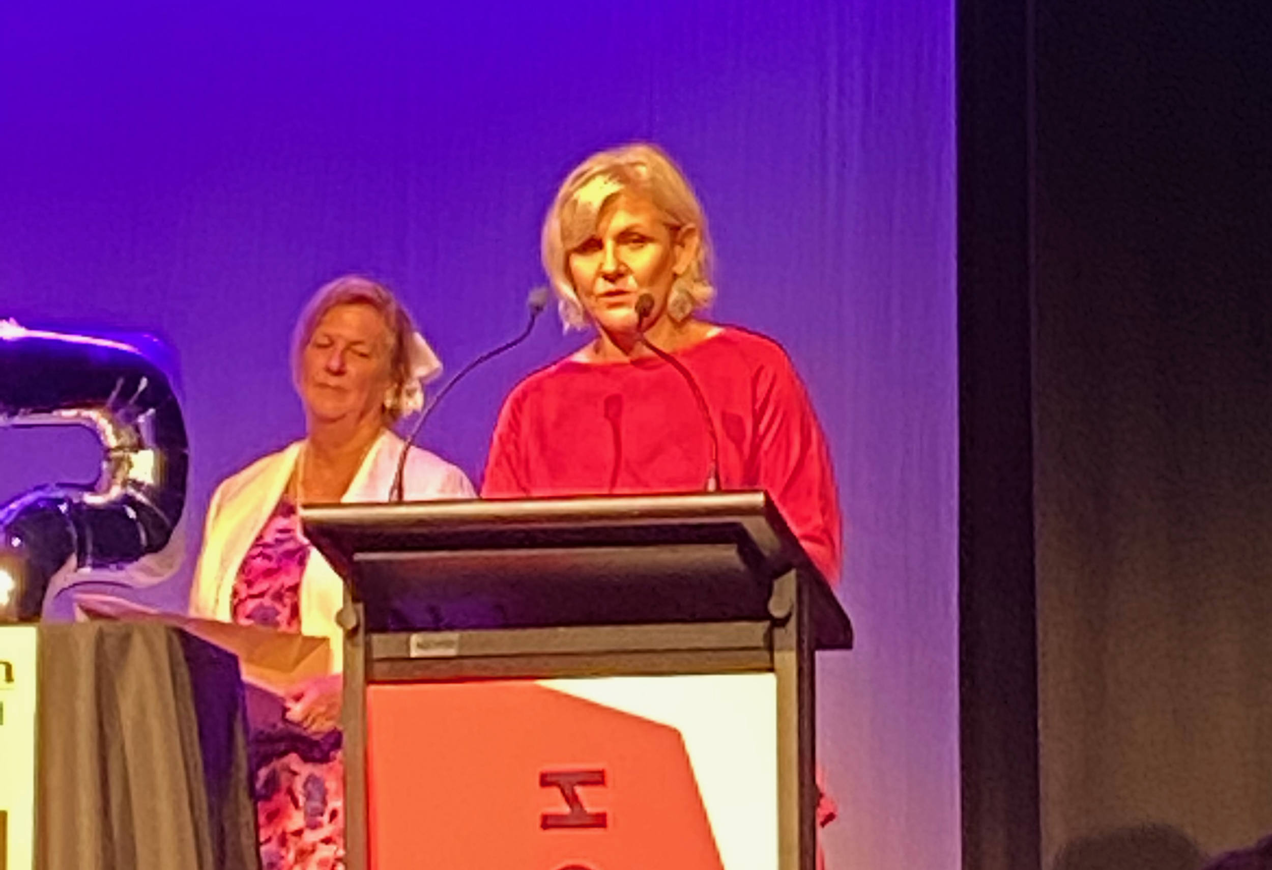 Two women onstage behind a podium under purple lighting, with a projected presentation screen in the background.