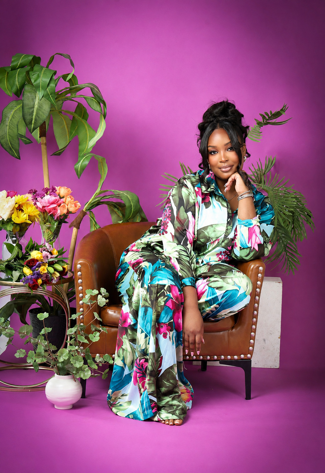 A woman sitting on a brown leather chair surrounded by tropical plants and colorful flowers, with a purple background, wearing a matching tropical print outfit.
