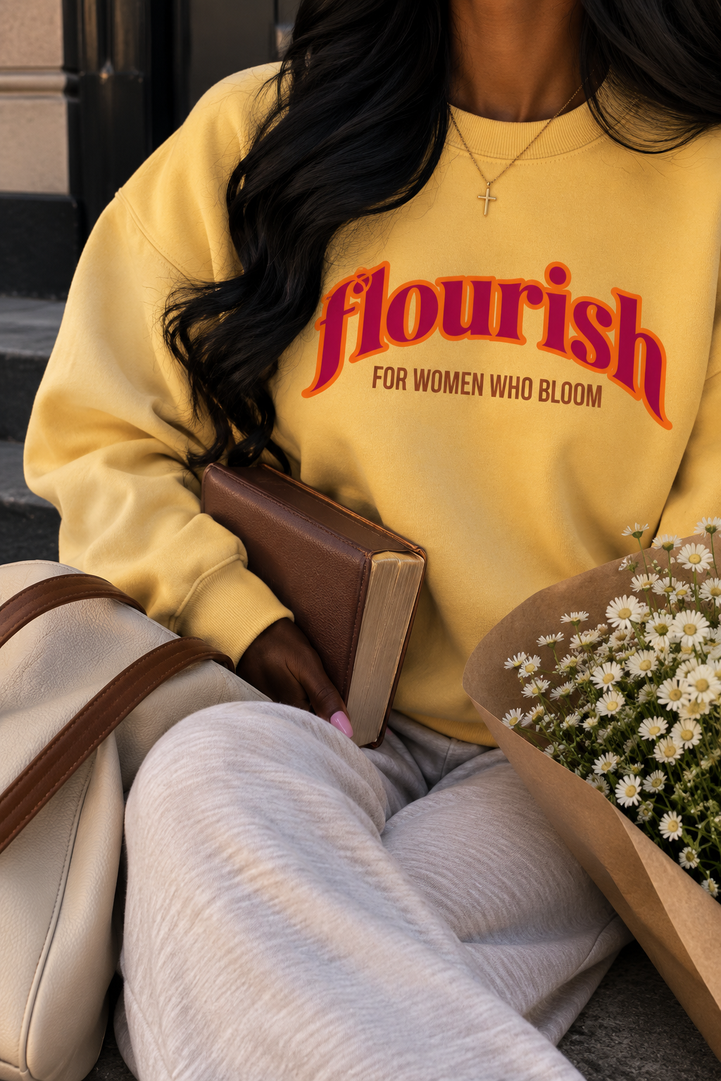 Close-up of a woman wearing a yellow sweatshirt with the word 'flourish' and the phrase 'for women who bloom,' holding a bouquet of daisies and a bible, with a beige purse and a brown bag nearby.